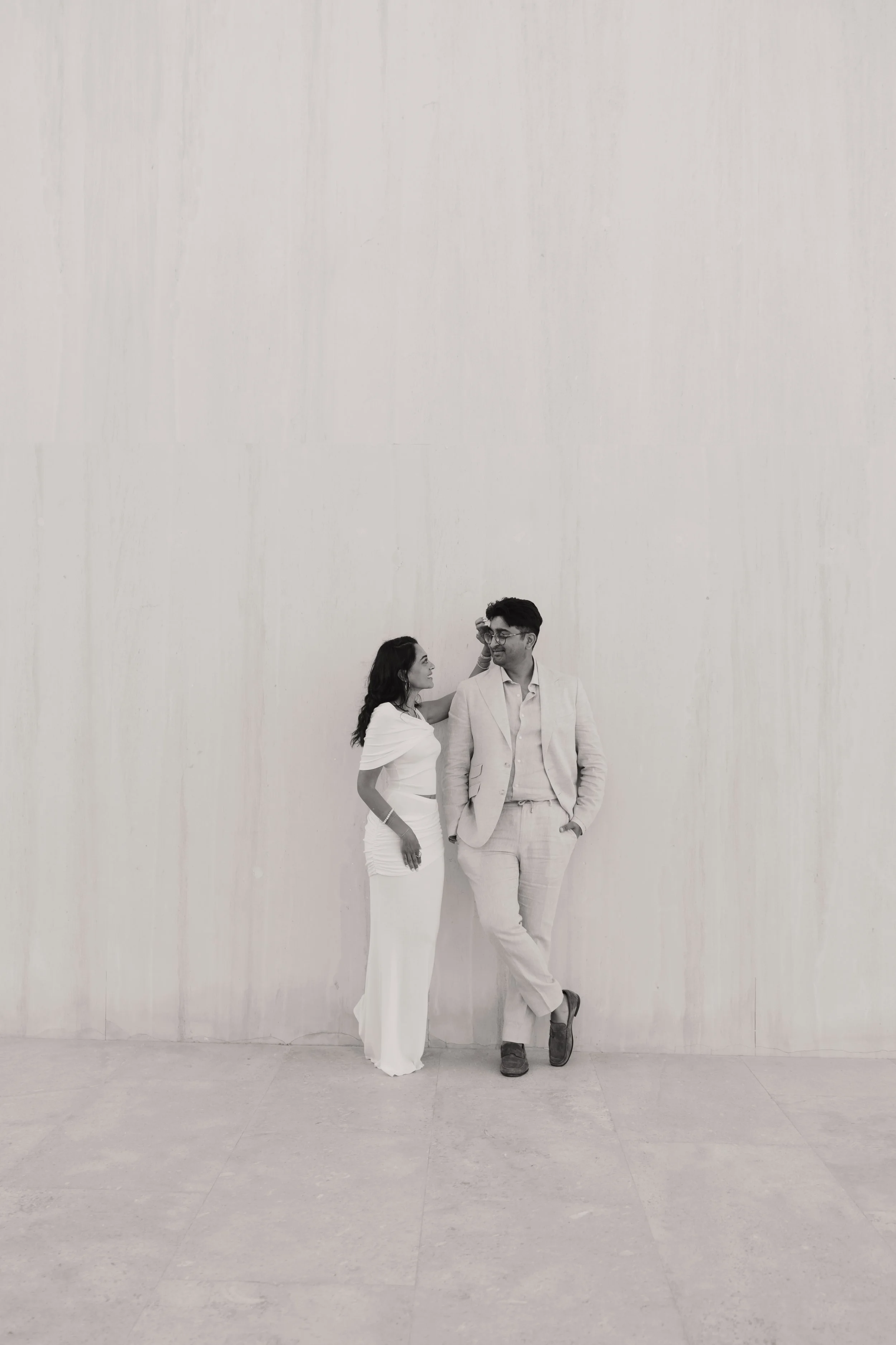 A black-and-white photo of a woman and a man standing against a plain wall. The woman is dressed in a white outfit with a cape, and the man is in a light-colored suit with glasses. The woman is touching the man's face while looking at him, and the ma