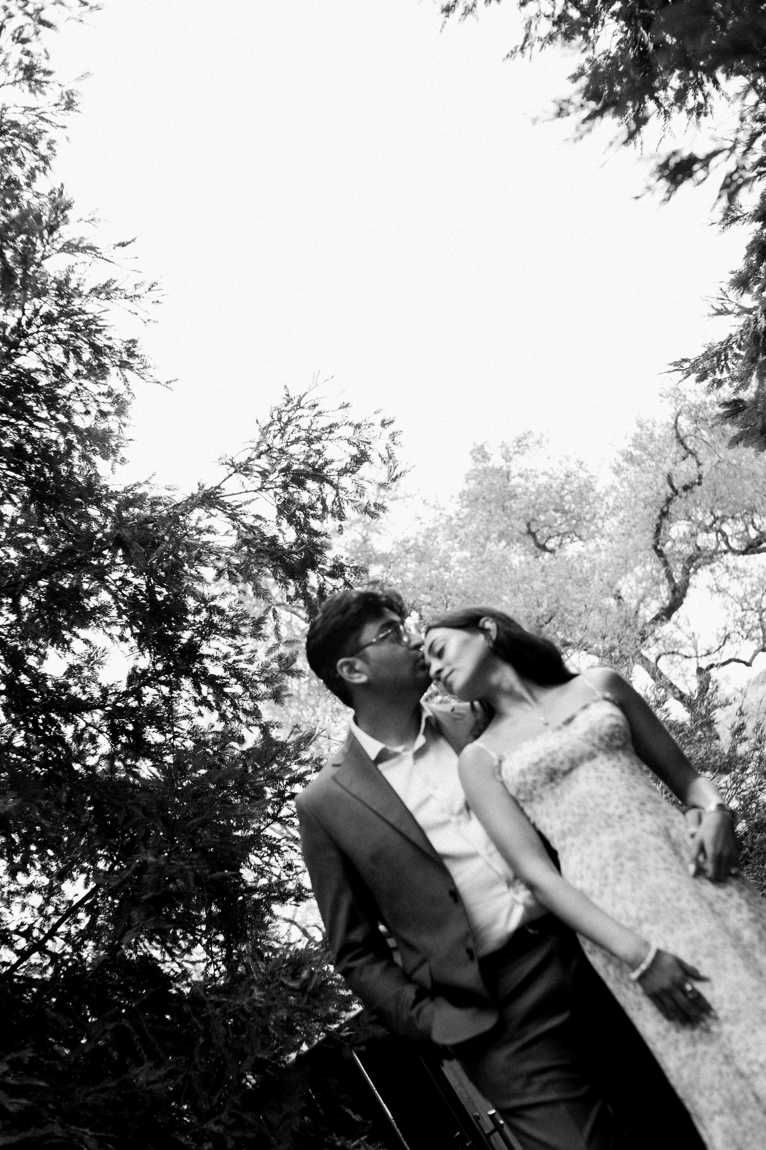 A black and white photo of a man and woman standing close together outdoors among trees, with the man kissing the woman on the forehead.