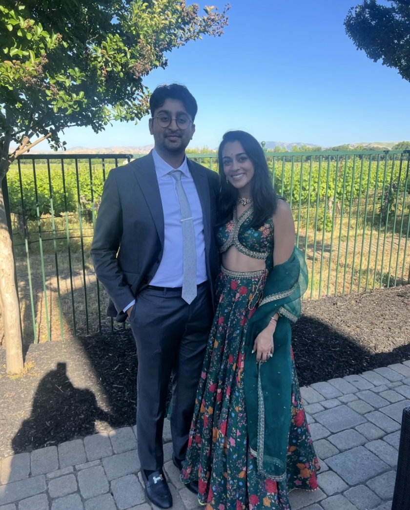 A man in a gray suit and tie standing next to a woman in a teal and floral traditional outfit, outdoors on a paved area with green vineyard and trees in the background under a clear blue sky.