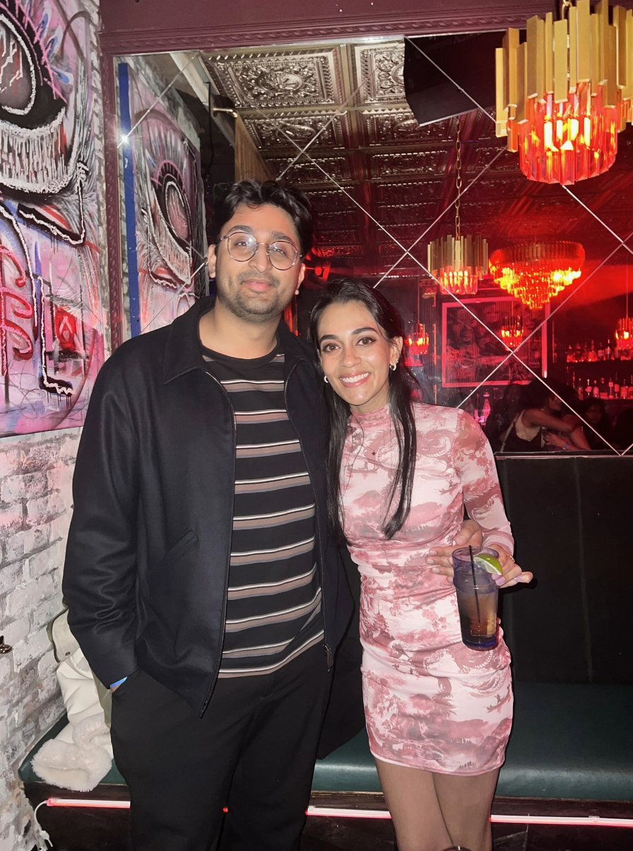 A man and woman posing together in a dimly lit bar or nightclub with red lighting, modern chandeliers, and graffiti art on the wall.
