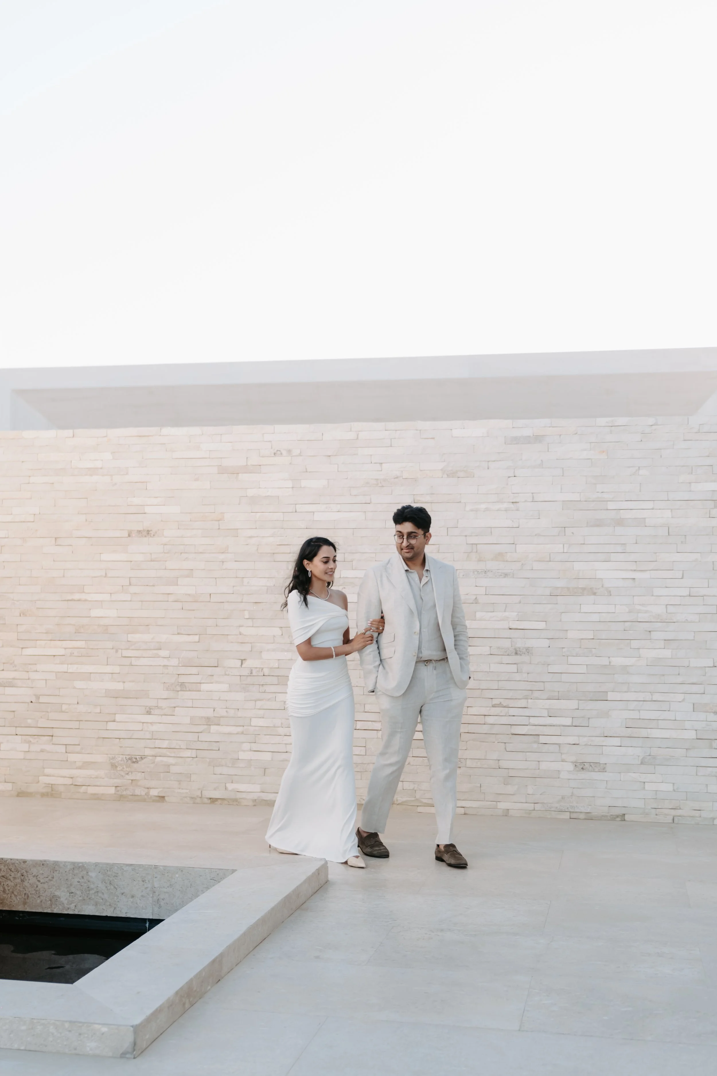 A couple dressed in white and beige suits standing outdoors in front of a light-colored brick wall, with a small square water feature nearby.