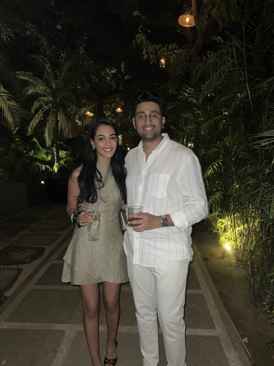 A woman and a man standing together outdoors at night, smiling and holding drinks, surrounded by lush greenery and hanging lanterns.