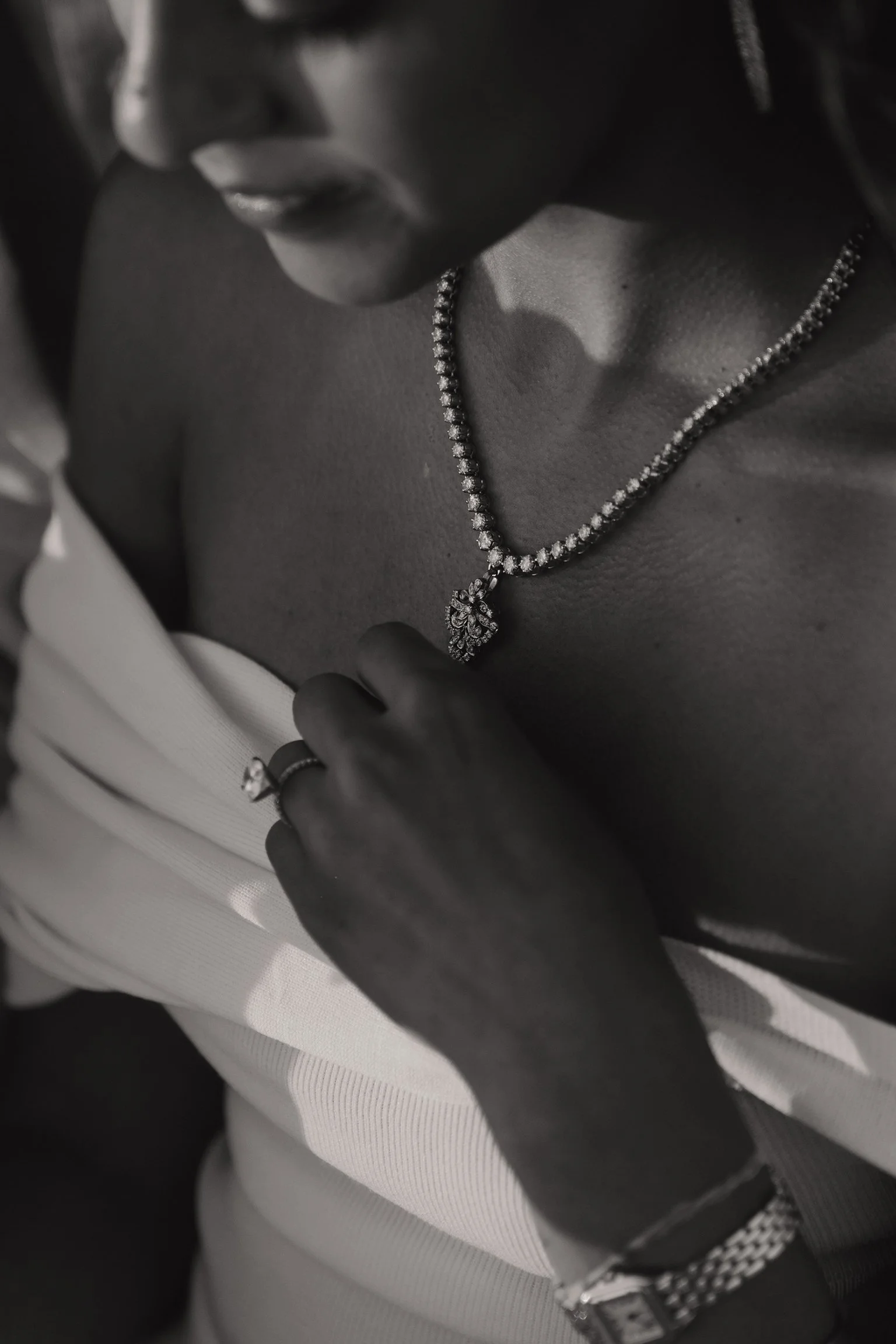 A person wearing a silver necklace with a pendant, a ring, and a watch. The person is wearing a light-colored top, and their hand is resting on their chest.