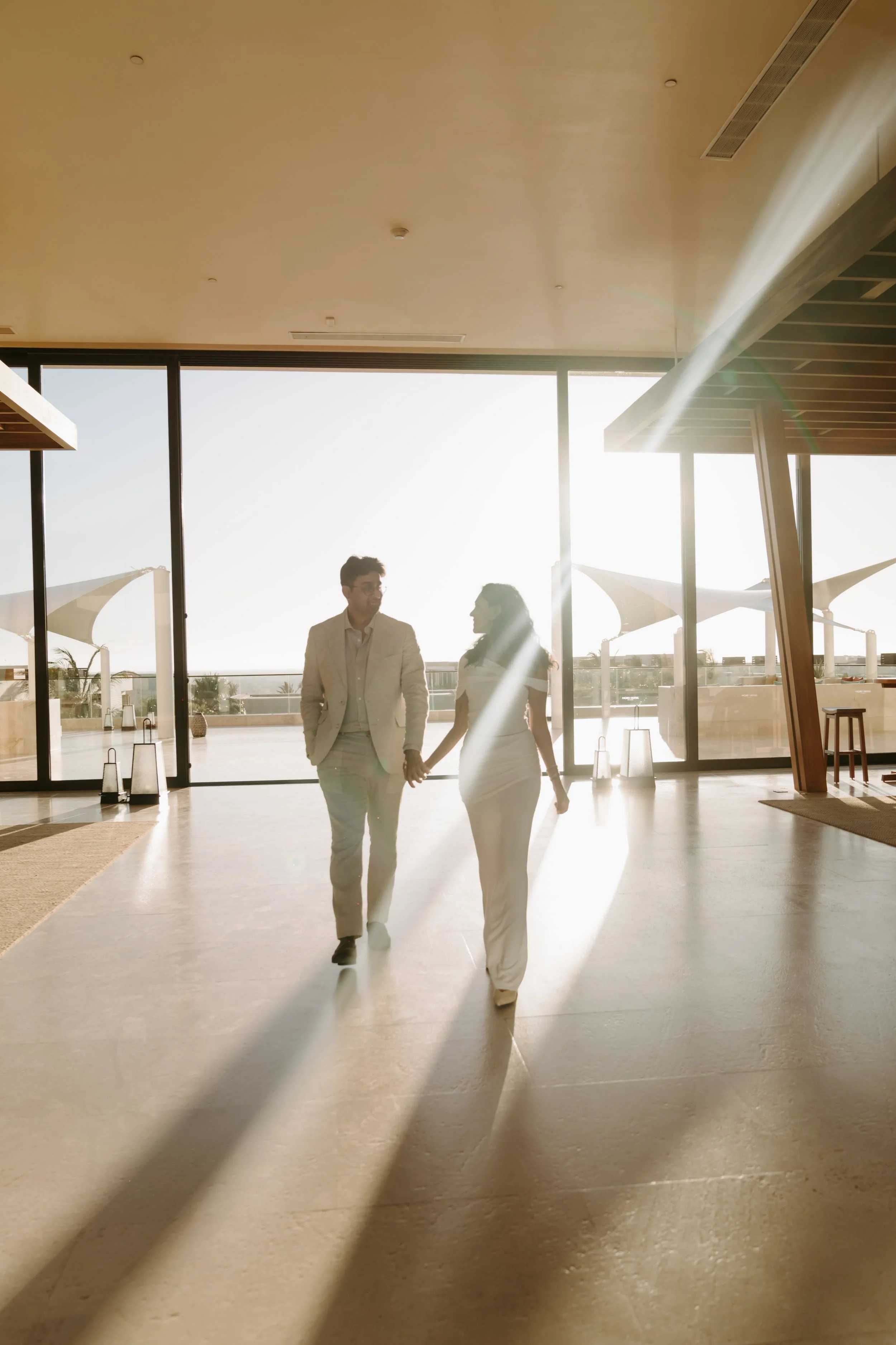 A couple holding hands walking inside a modern building with large glass windows and sunlight streaming in.