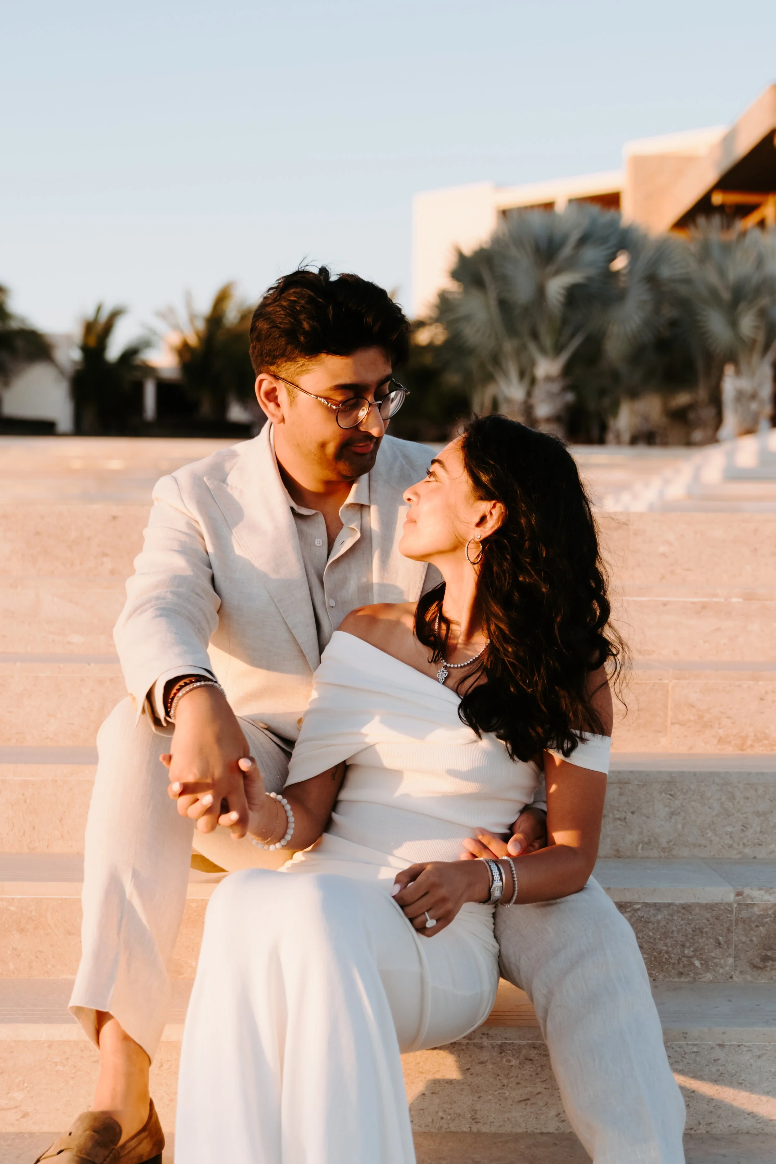A couple sitting on steps outdoors during sunset, gazing at each other affectionately. The man is wearing glasses, a light-colored suit, and the woman is in a white off-shoulder dress.