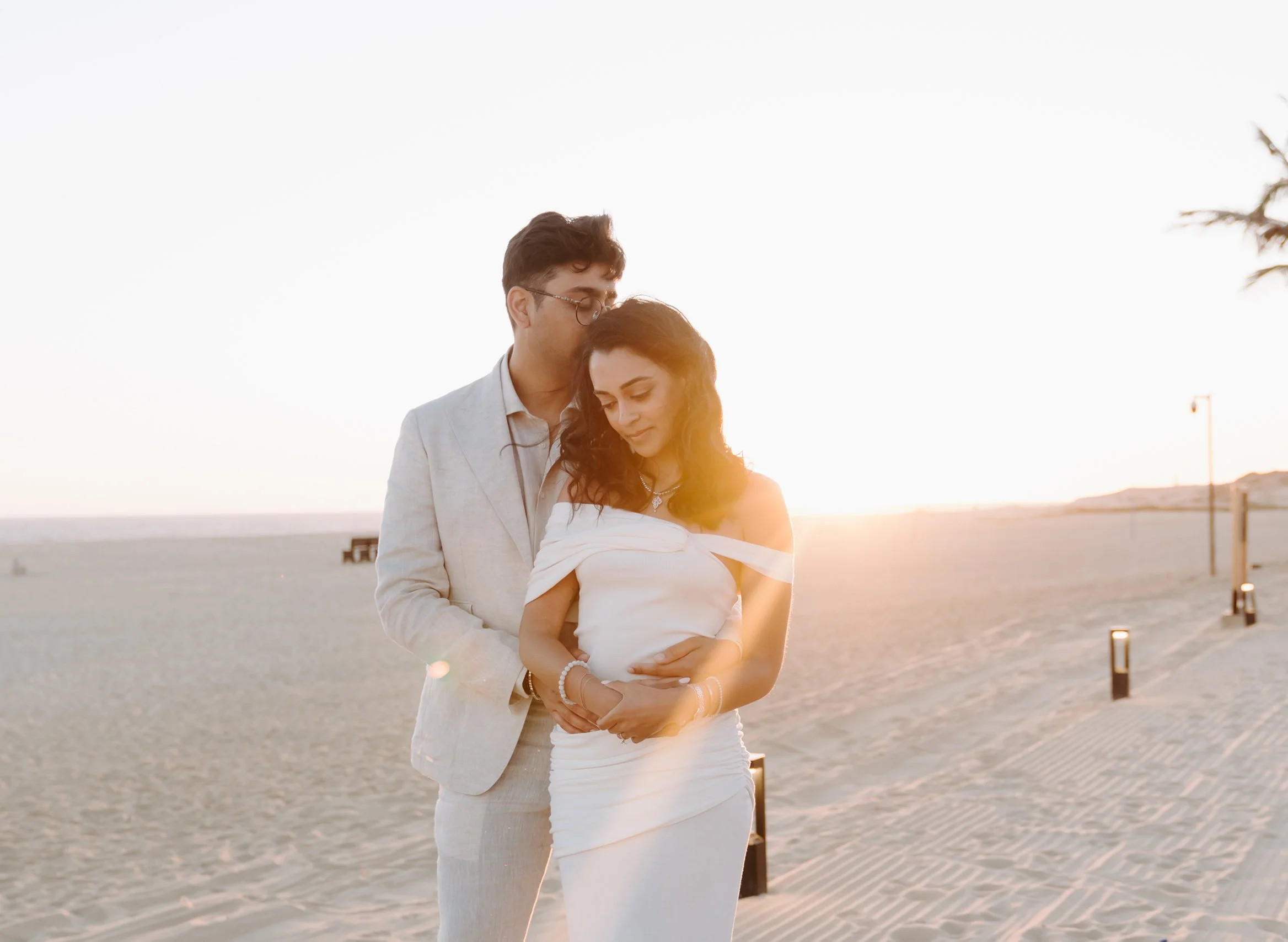 A couple dressed in wedding attire standing on a beach at sunset, with the man kissing the woman's forehead and the woman smiling with eyes closed.
