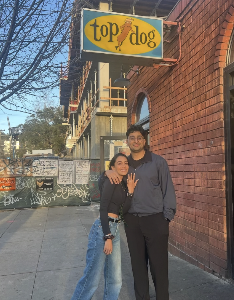 Two people standing outside a restaurant named Top Dog, with a bright yellow sign and a cartoon hot dog wearing a chef's hat. One person has their arm around the other and is showing off a ring.