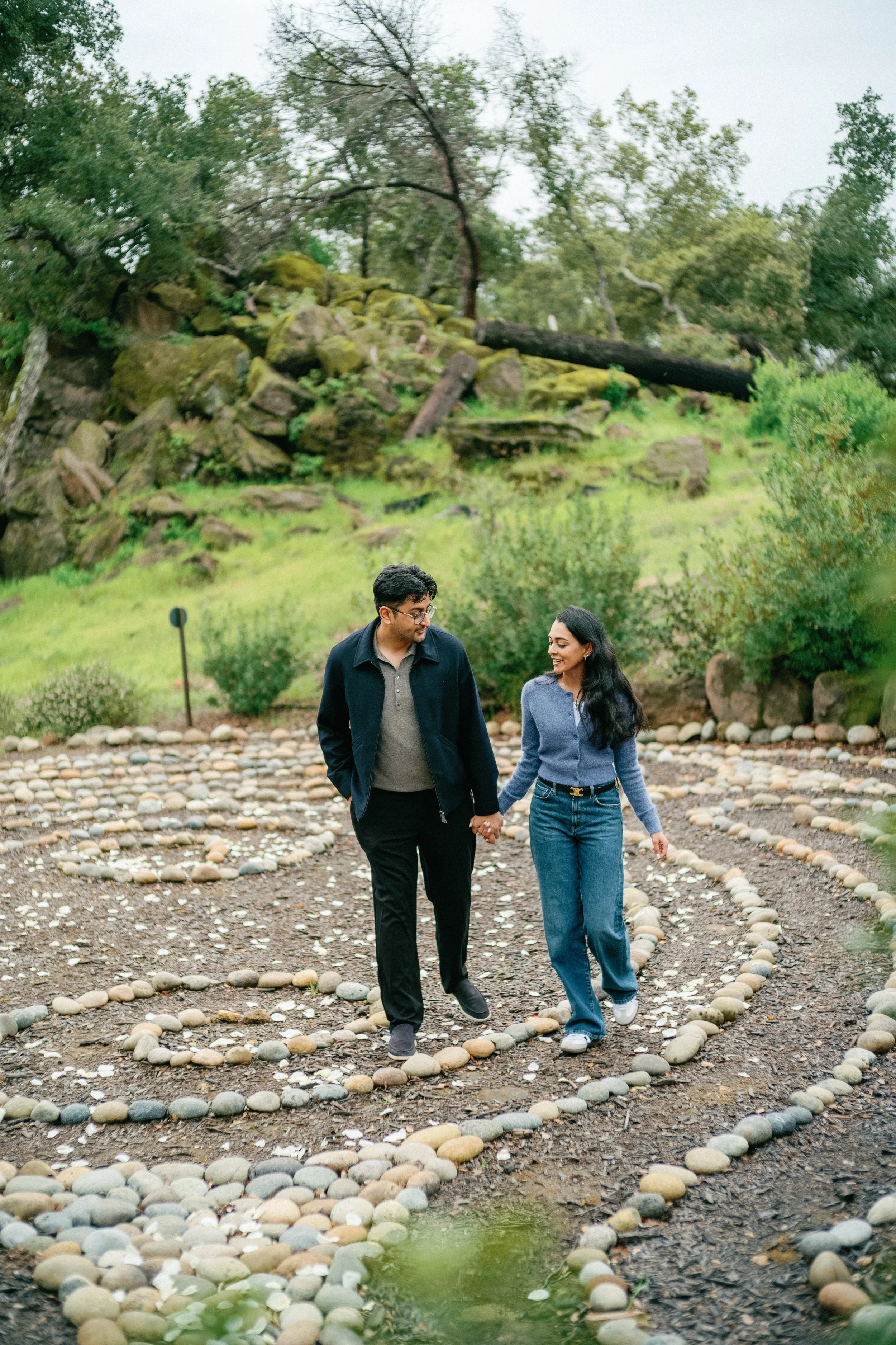 A couple holding hands and walking along a pebble-lined path outdoors, with greenery and trees in the background.
