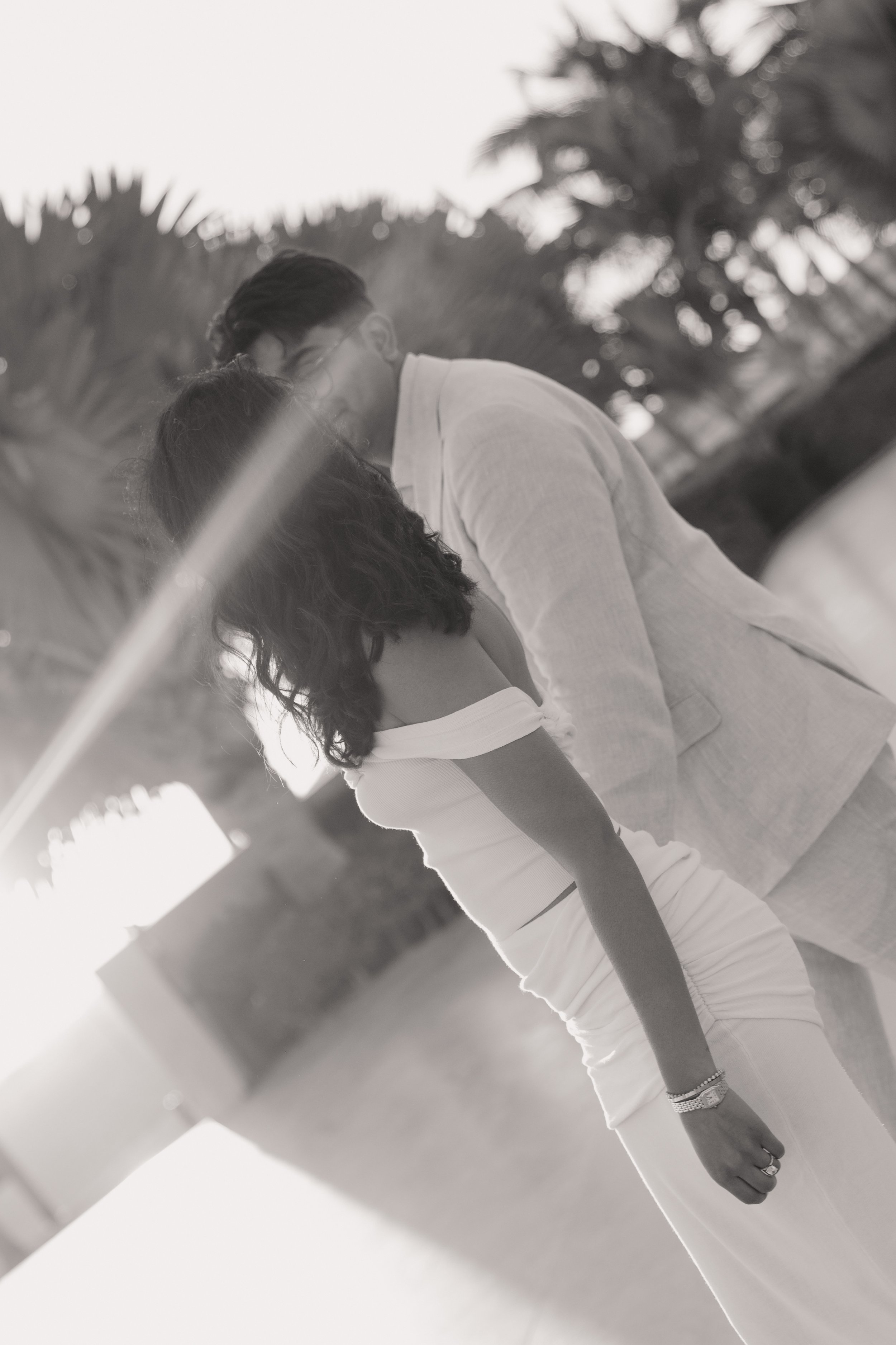 A black and white photo of a couple standing close together outdoors amidst palm trees, with the sun setting in the background.
