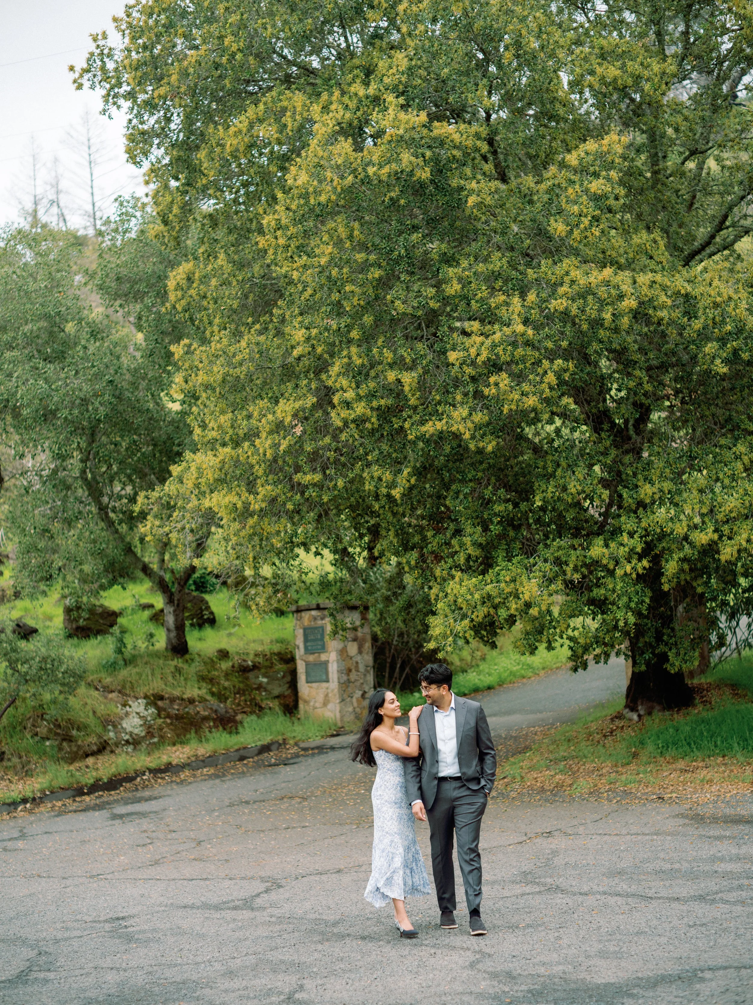 A couple walking arm in arm on a paved road beneath a large leafy tree in a park with green grass and smaller trees in the background.