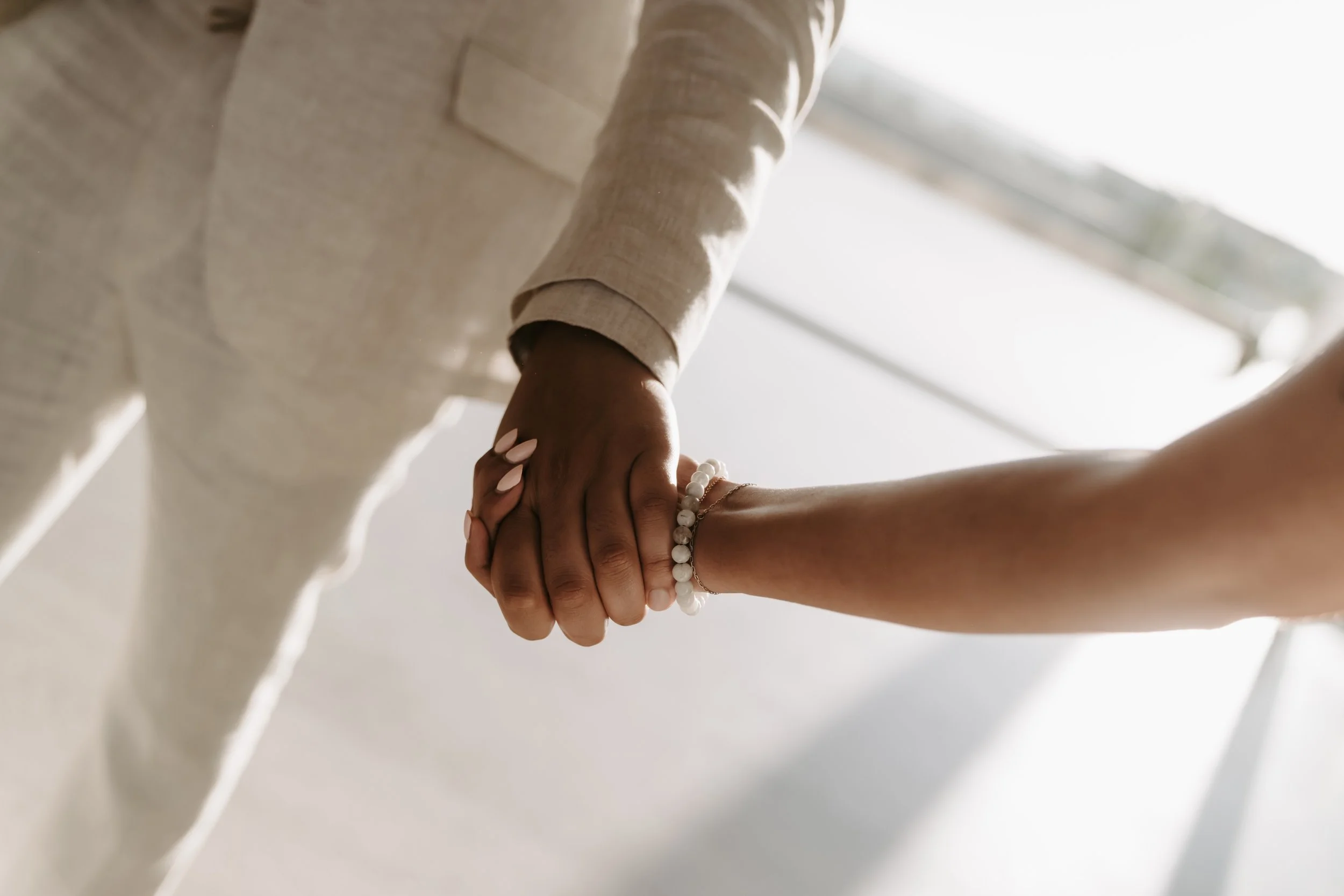 Two people holding hands, one with darker skin wearing a white long-sleeve shirt and the other with lighter skin wearing a bracelet, against a bright window background.
