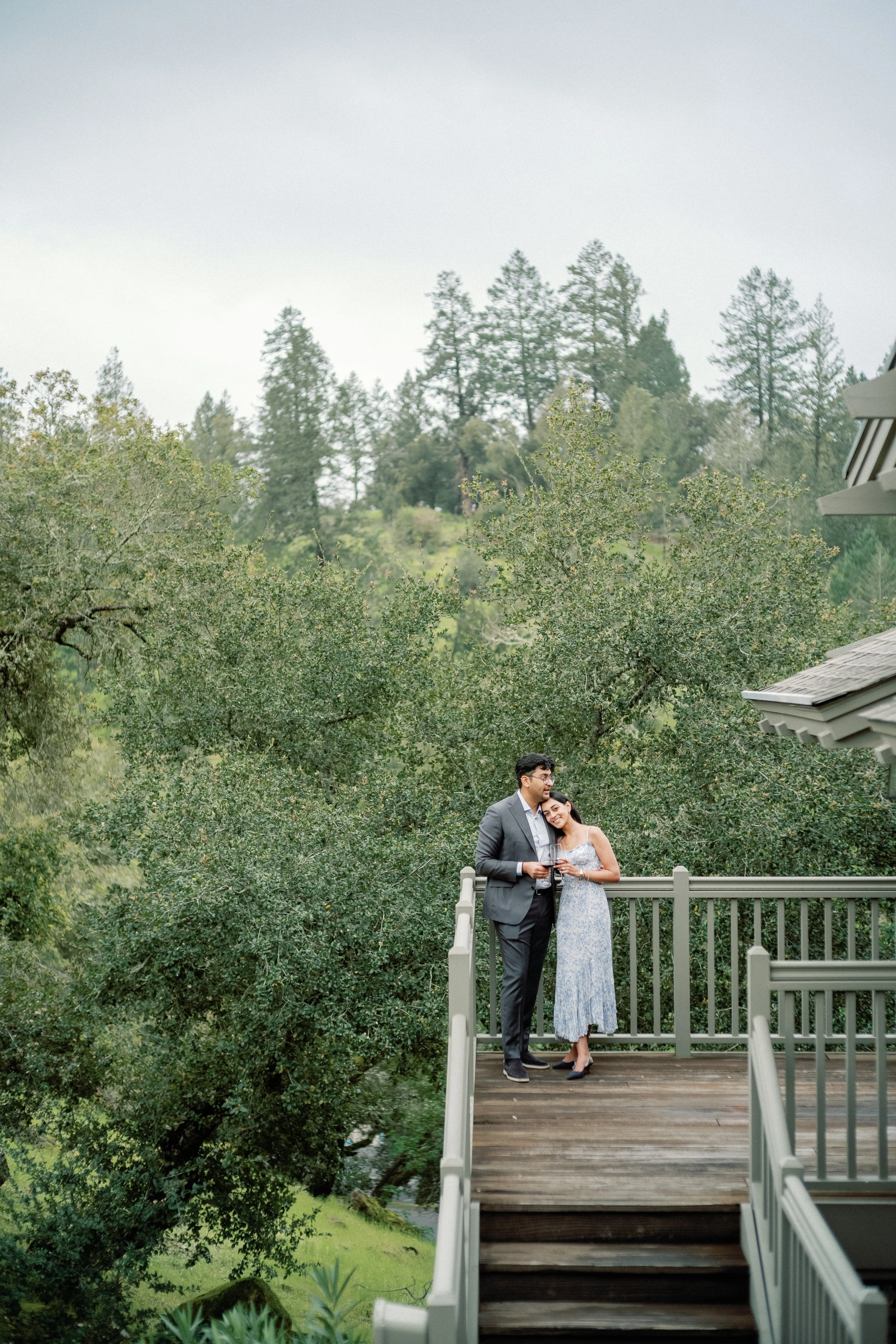 A couple standing on a balcony holding glasses of wine, surrounded by trees and a natural landscape.