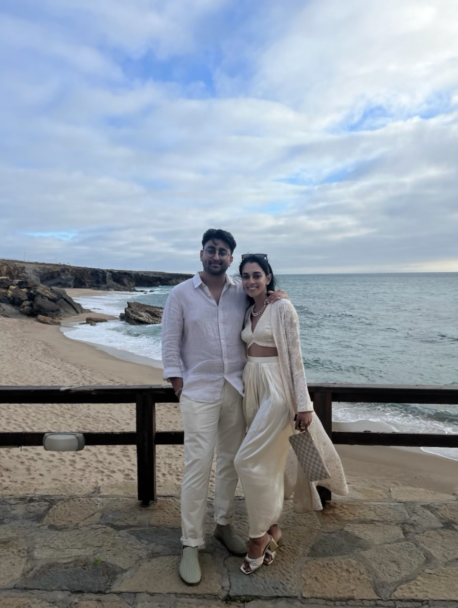 A smiling couple stands by a beach with rocks and the ocean in the background, under a partly cloudy sky.