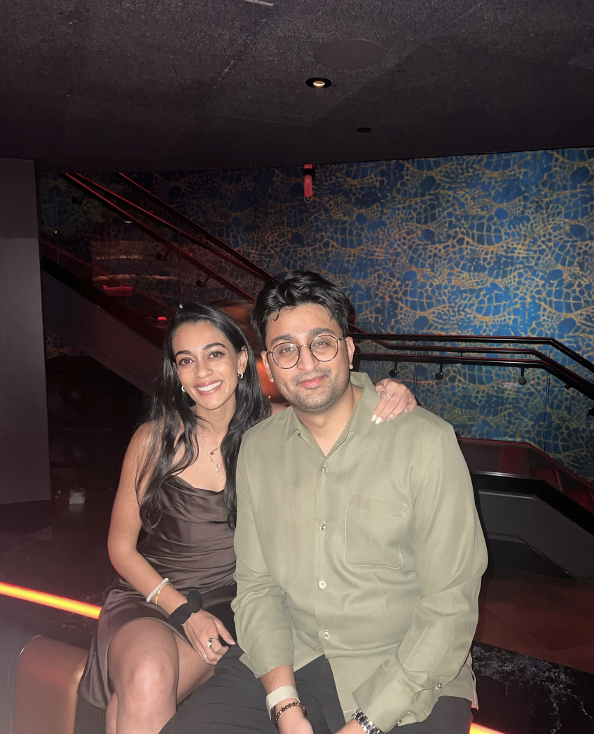 A man and a woman sitting together in a dimly lit room, smiling at the camera. The woman has long dark hair and is wearing a dark dress, while the man has short dark hair, glasses, and is wearing a light green shirt. They are seated in front of a sta