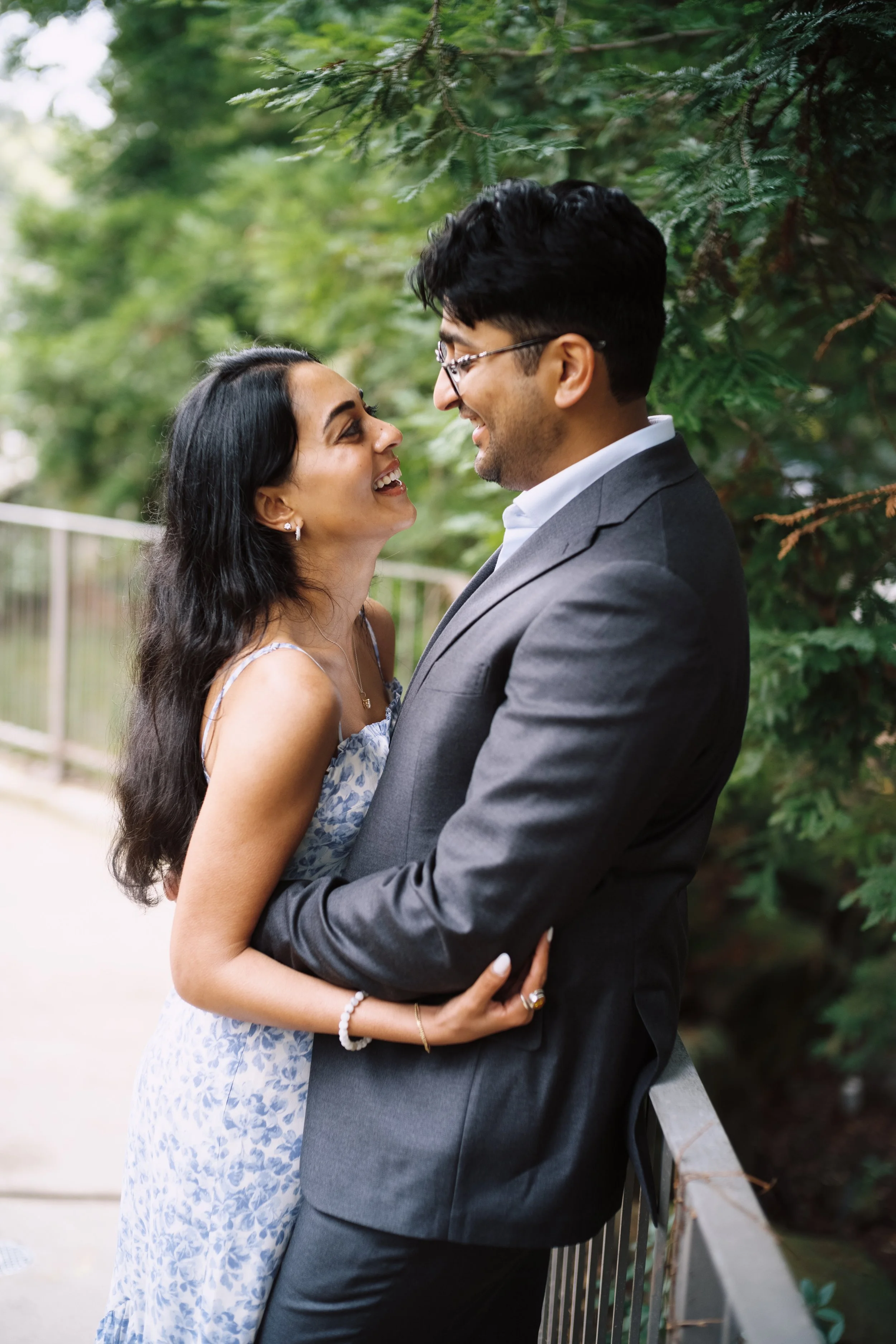A couple standing close together outdoors, smiling and looking into each other's eyes, with green trees in the background.