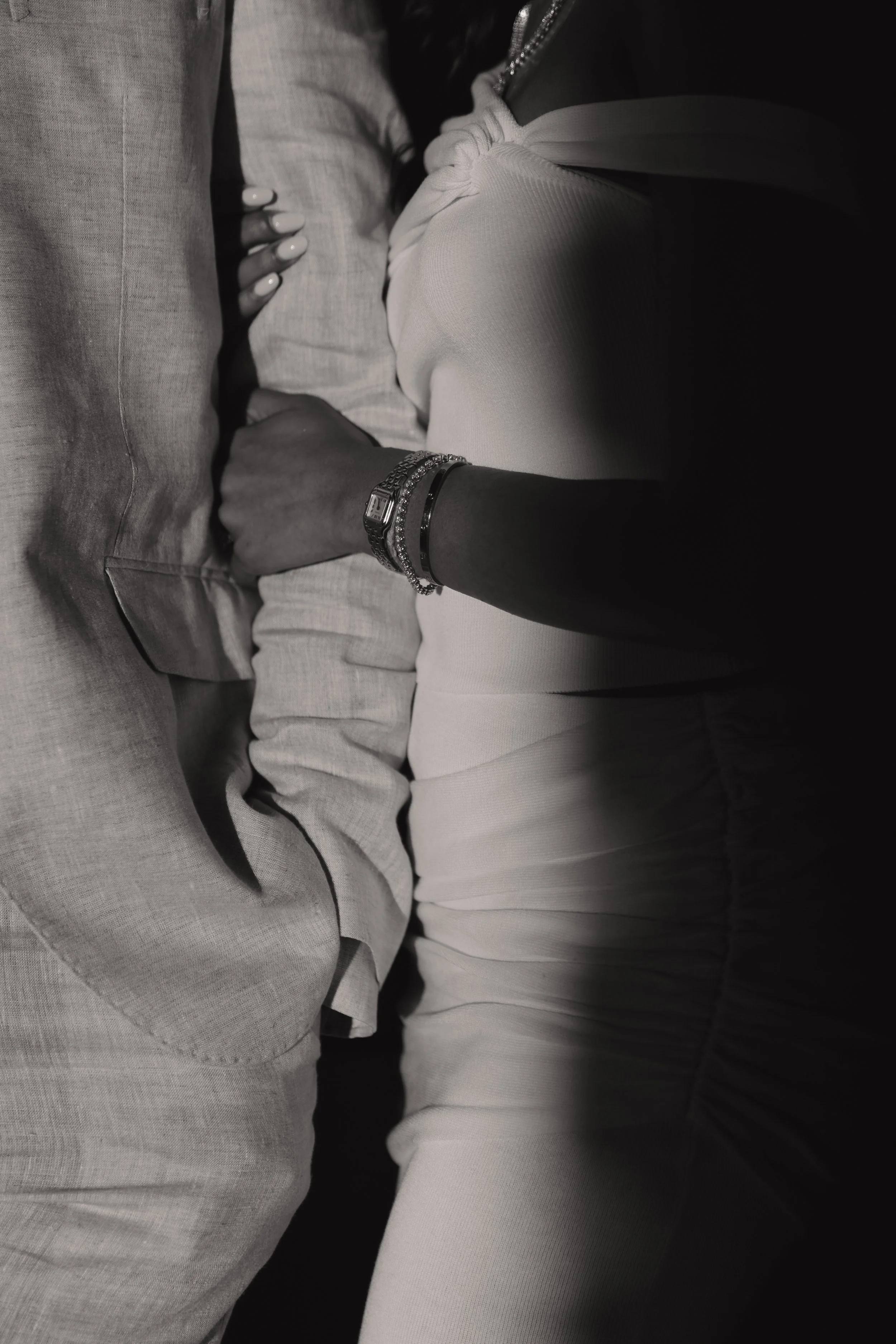 Close-up of two people holding hands, one wearing a watch and bracelets, their arms intertwined, in black and white.