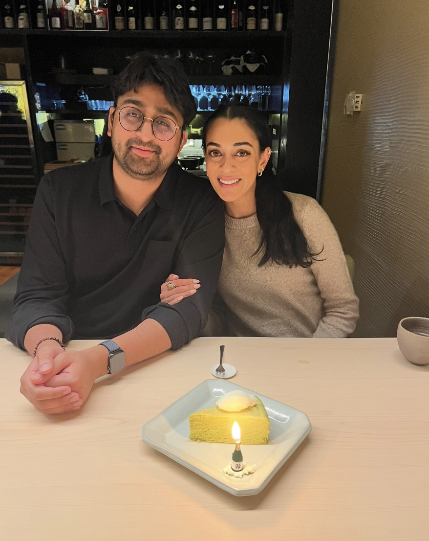 A man and a woman sit at a restaurant table celebrating with a slice of yellow cake with a lit candle and a scoop of ice cream on top. The background shows a bar with bottles and glasses.