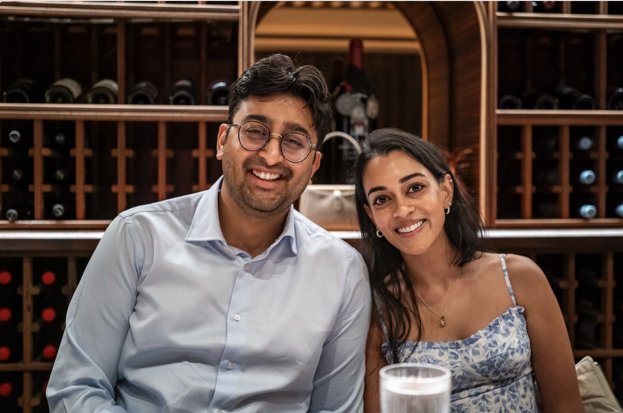 A smiling man with glasses and a woman sitting together at a table inside a wine cellar, with wine bottles stored in wooden racks in the background.