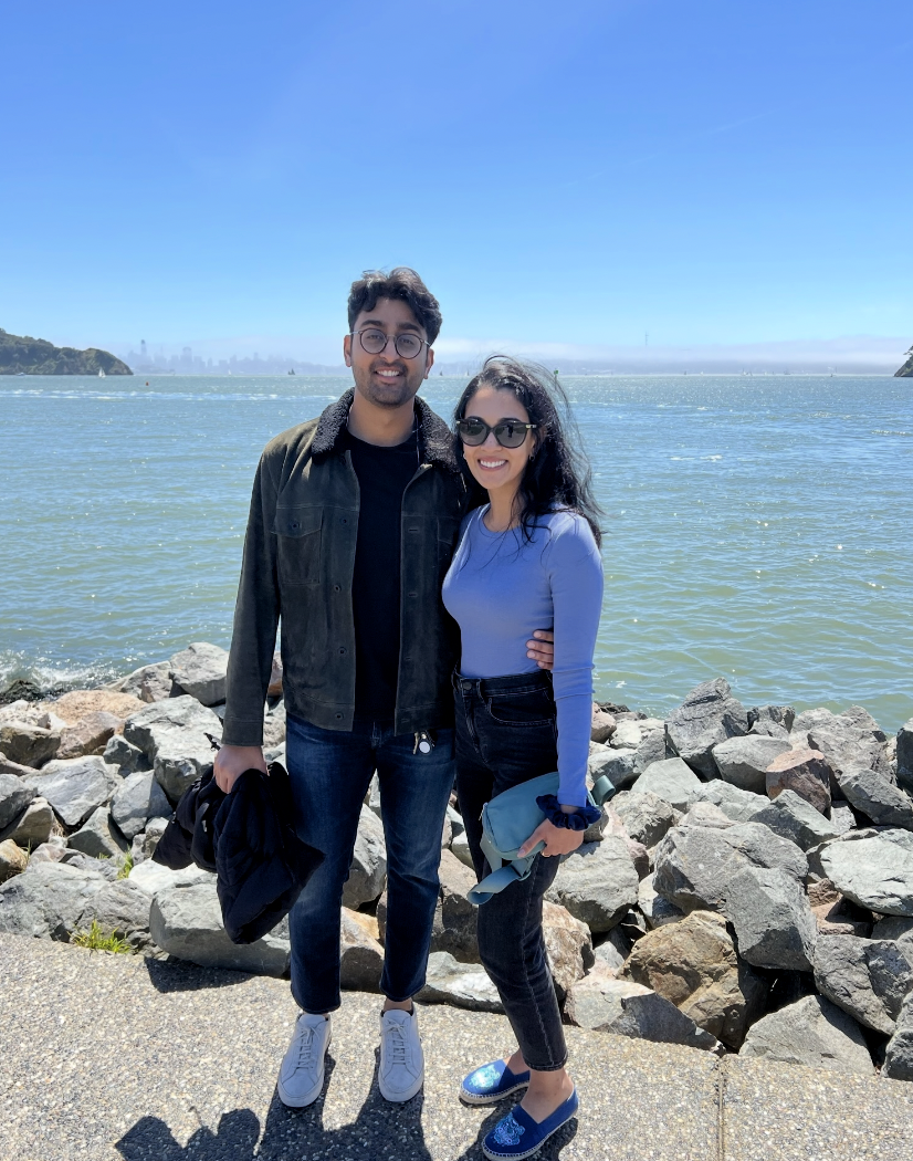 A man and woman standing on rocks near a body of water with a city skyline in the background, under a clear blue sky. They are smiling and dressed casually, with the woman wearing sunglasses and holding a rolled-up jacket.