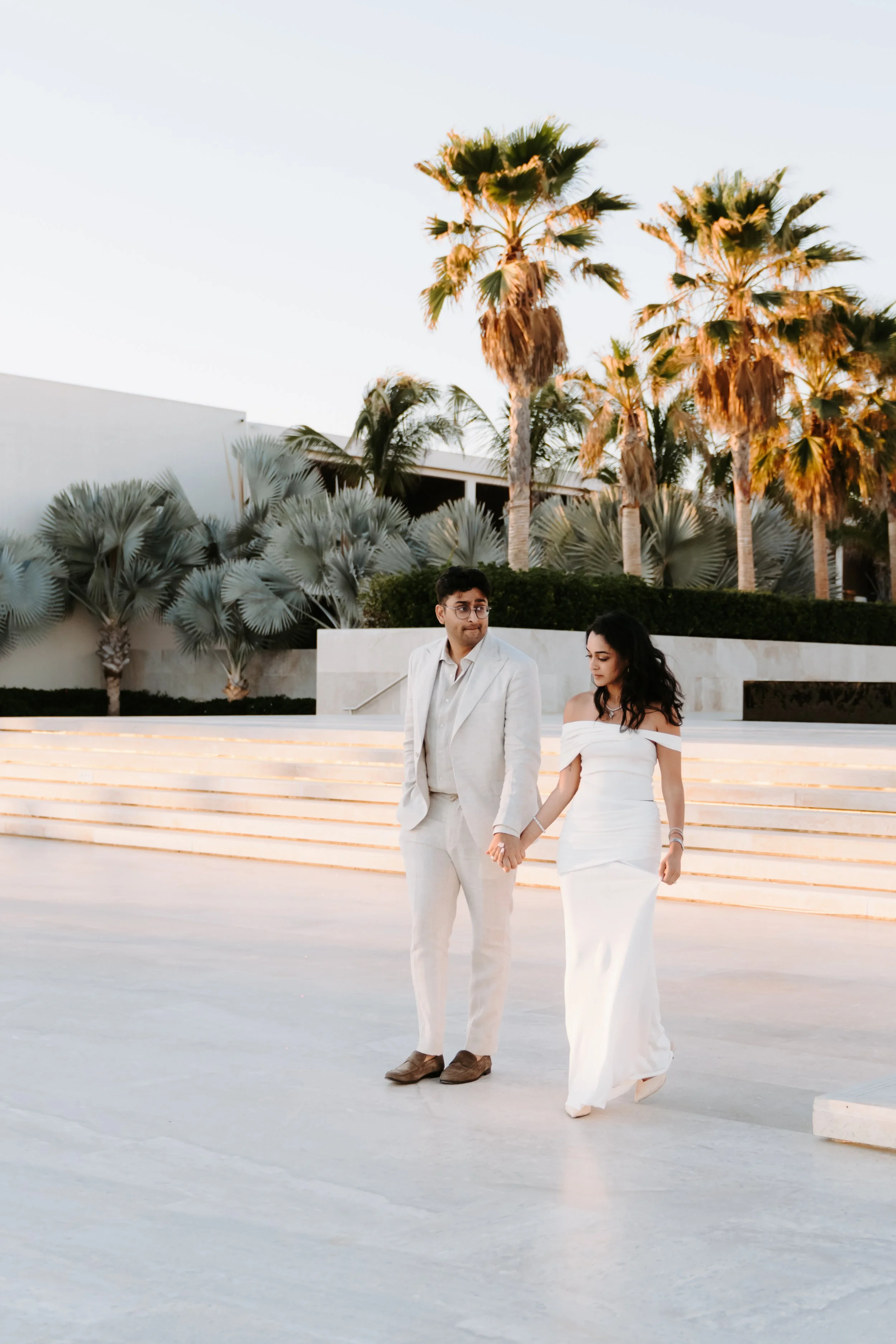 A man and woman in wedding attire walking hand in hand outdoors near palm trees at sunset.