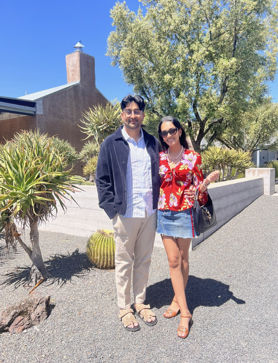 A man and a woman standing outdoors on a sunny day with desert plants and a large tree in the background. The man is wearing glasses, a dark jacket, beige pants, and sandals. The woman is dressed in a red floral top, denim skirt, sandals, and sunglas