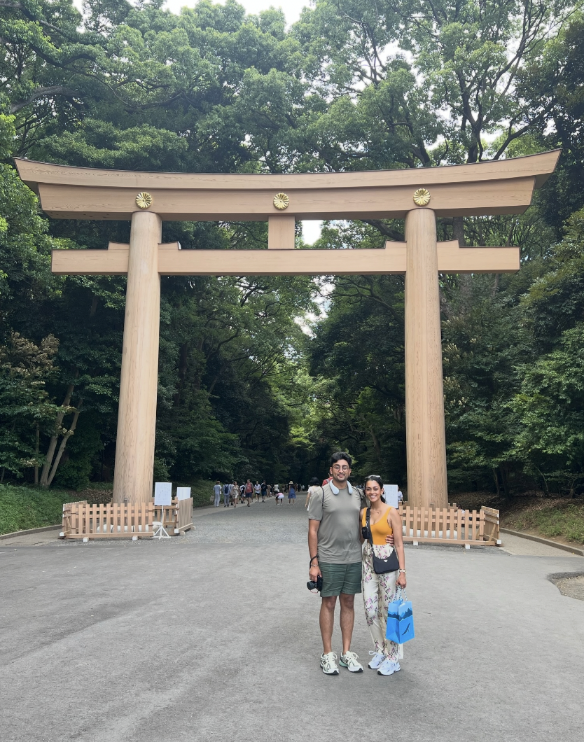 Two people standing in front of a large wooden torii gate at a lush green park or shrine.