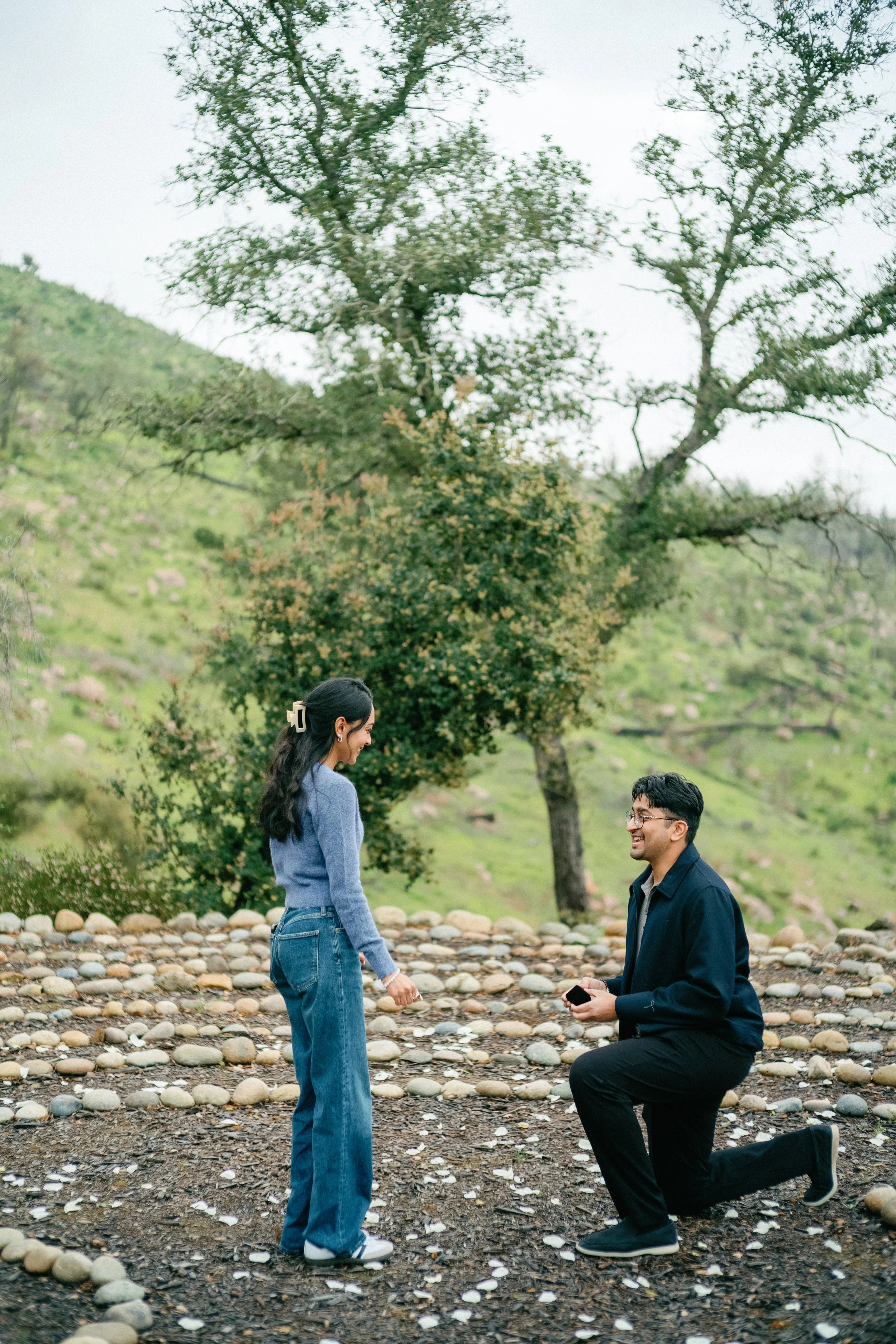 A man kneeling on one knee proposing to a woman outdoors on a rocky path with green trees in the background.
