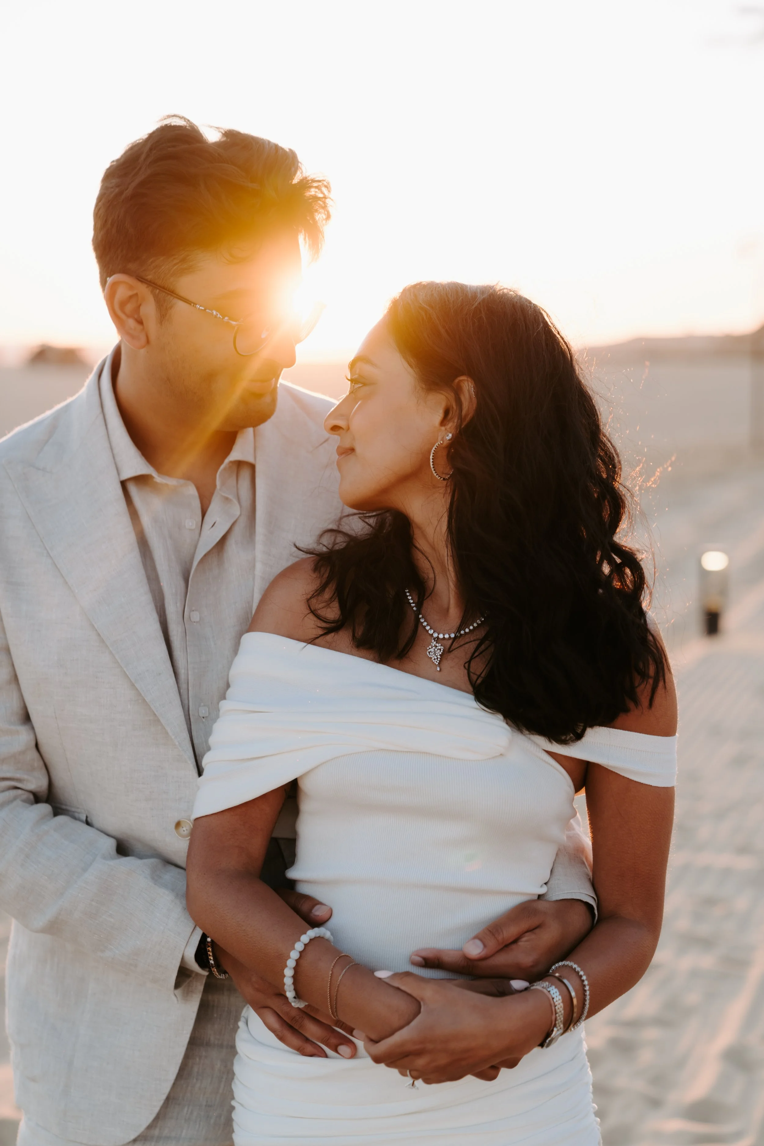 A couple standing close together on a beach at sunset, with the sun shining behind them.