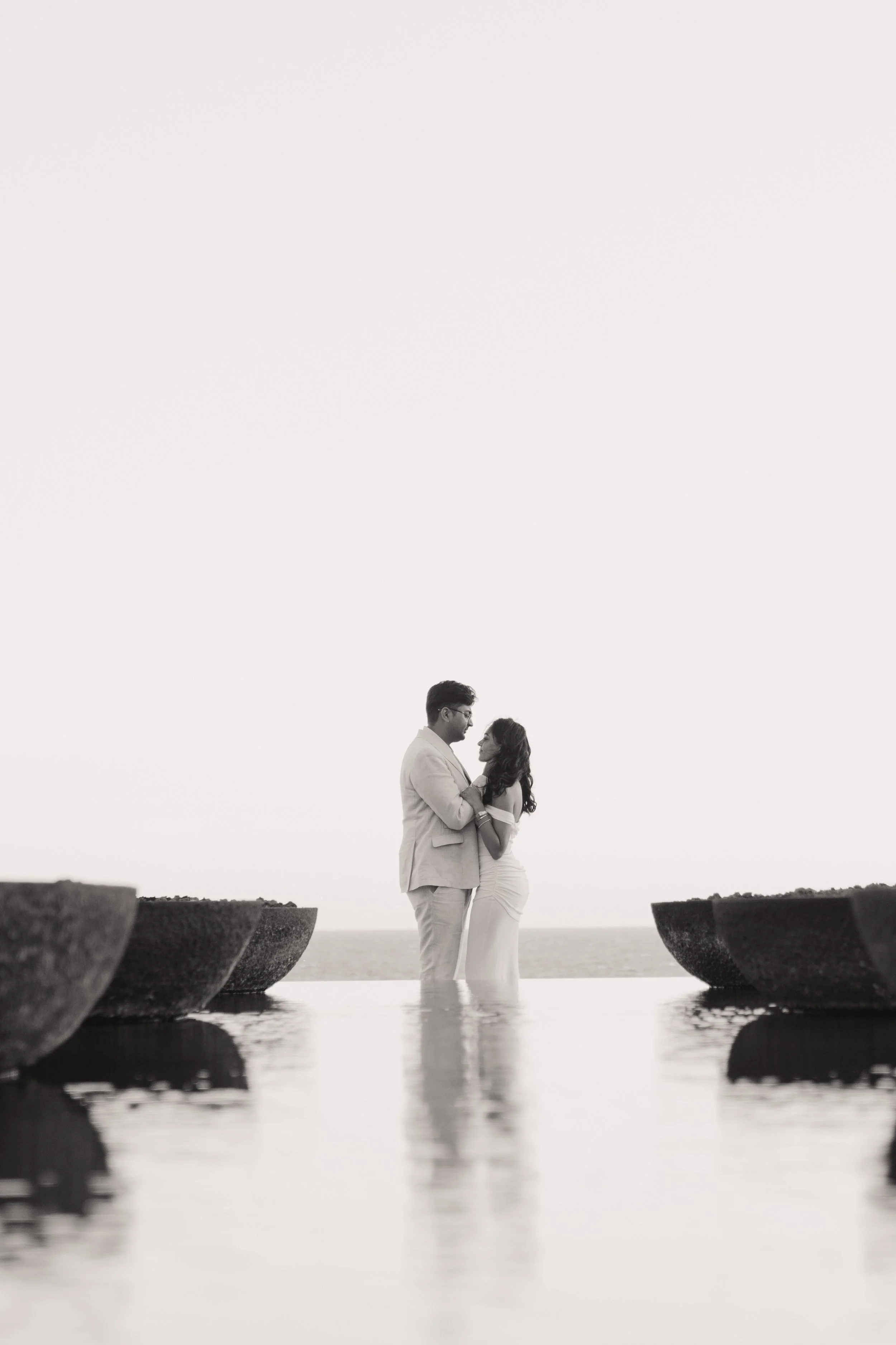A black and white photograph of a couple standing in water, facing each other and holding hands, with a clear sky and ocean in the background, framed by large stone structures.