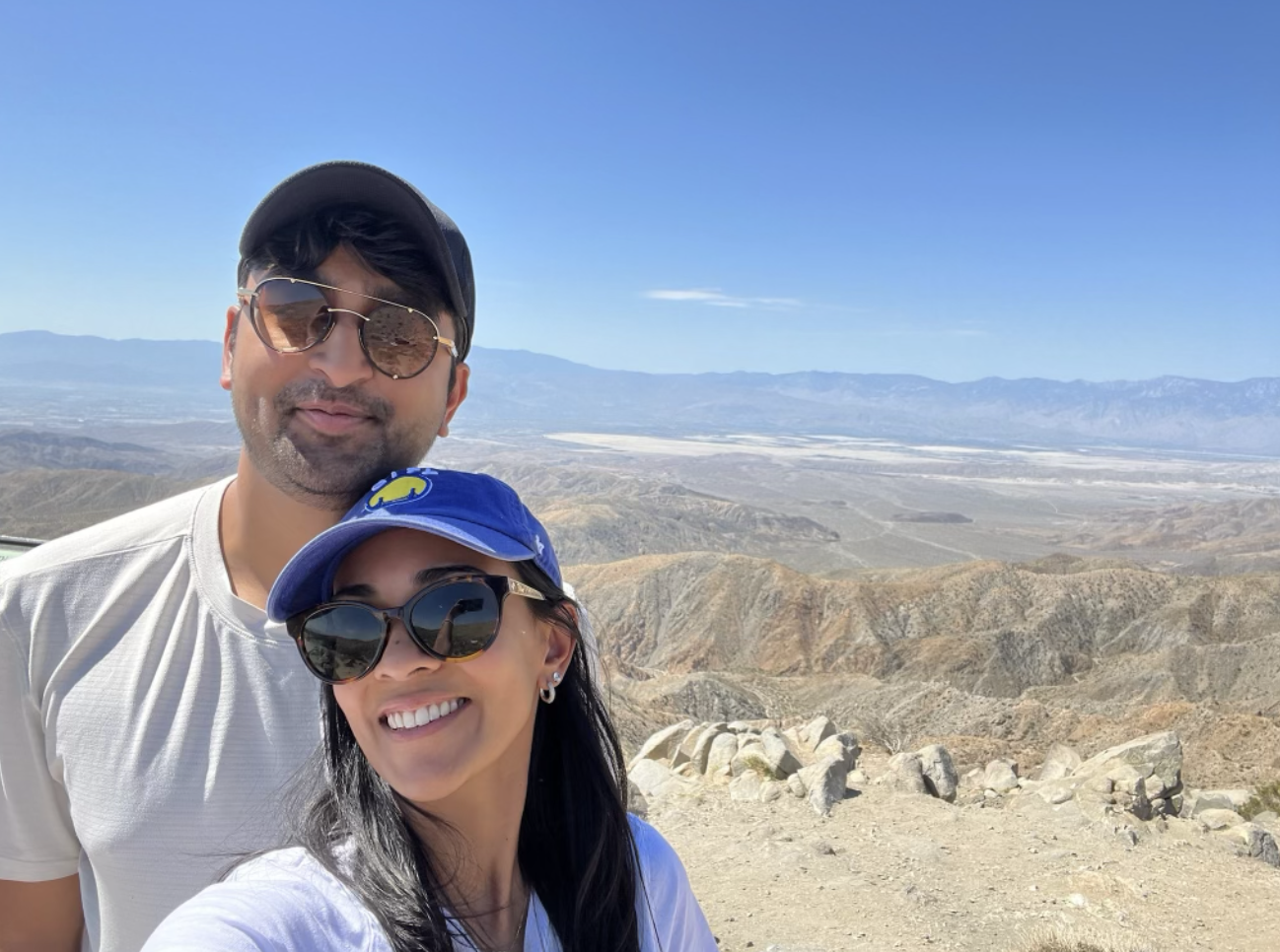 A smiling man and woman taking a selfie outdoors with a mountainous desert landscape in the background on a sunny day.