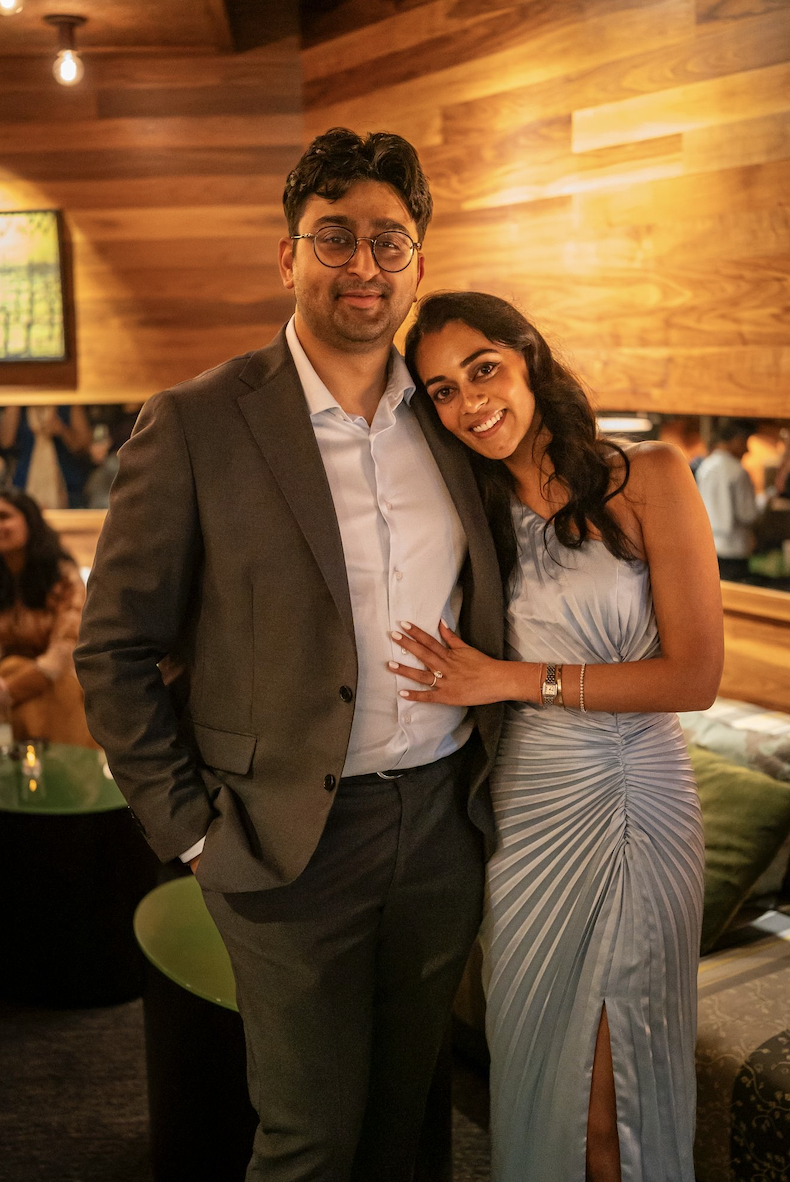 A man in a suit and a woman in a dress stand together, smiling, in a warmly lit, modern indoor setting with wooden wall paneling, other people in the background, and a television on the wall.