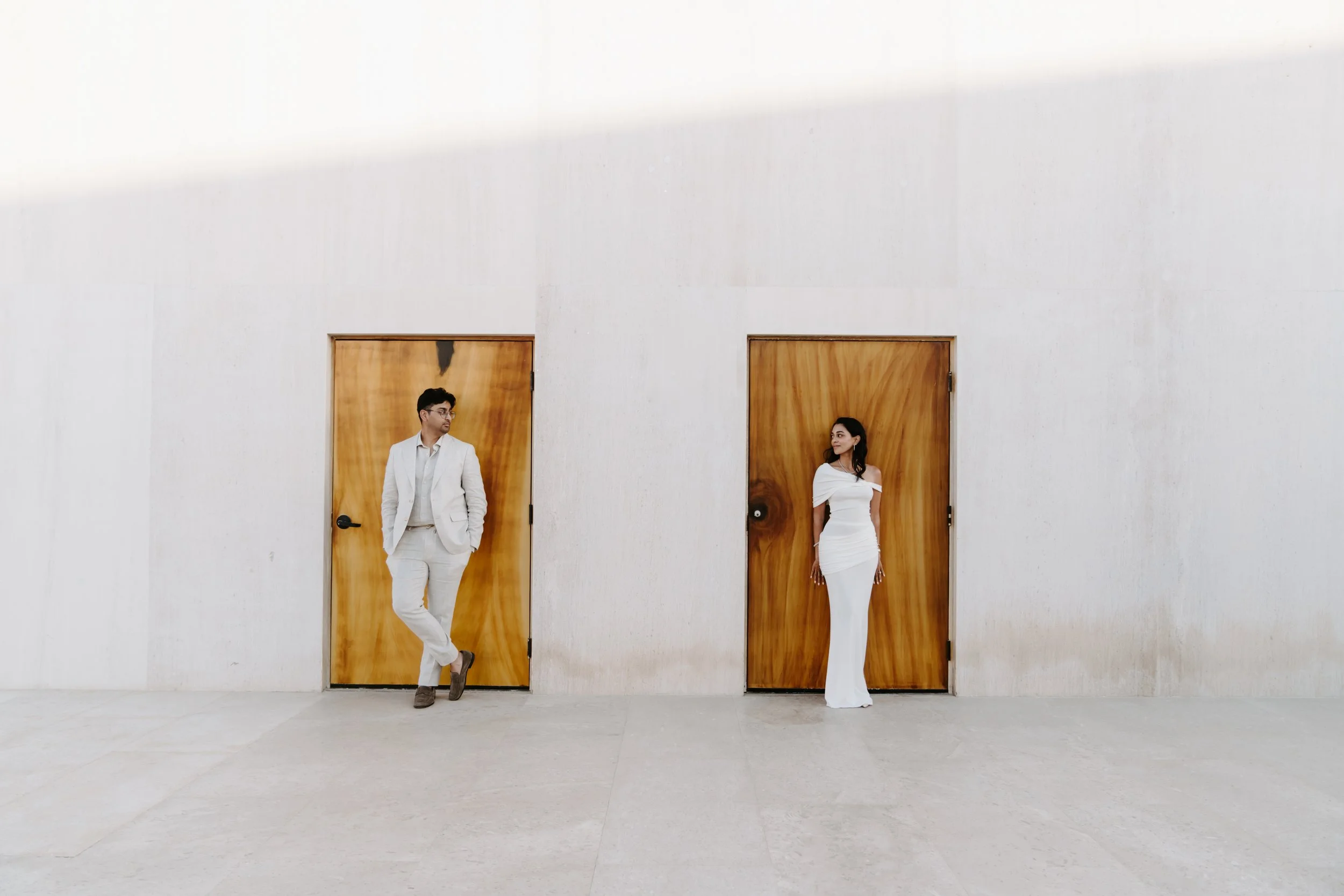 A man and woman standing apart in front of two wooden doors with white walls, the man leaning casually with his hands in his pockets and the woman standing with her arms relaxed at her sides.