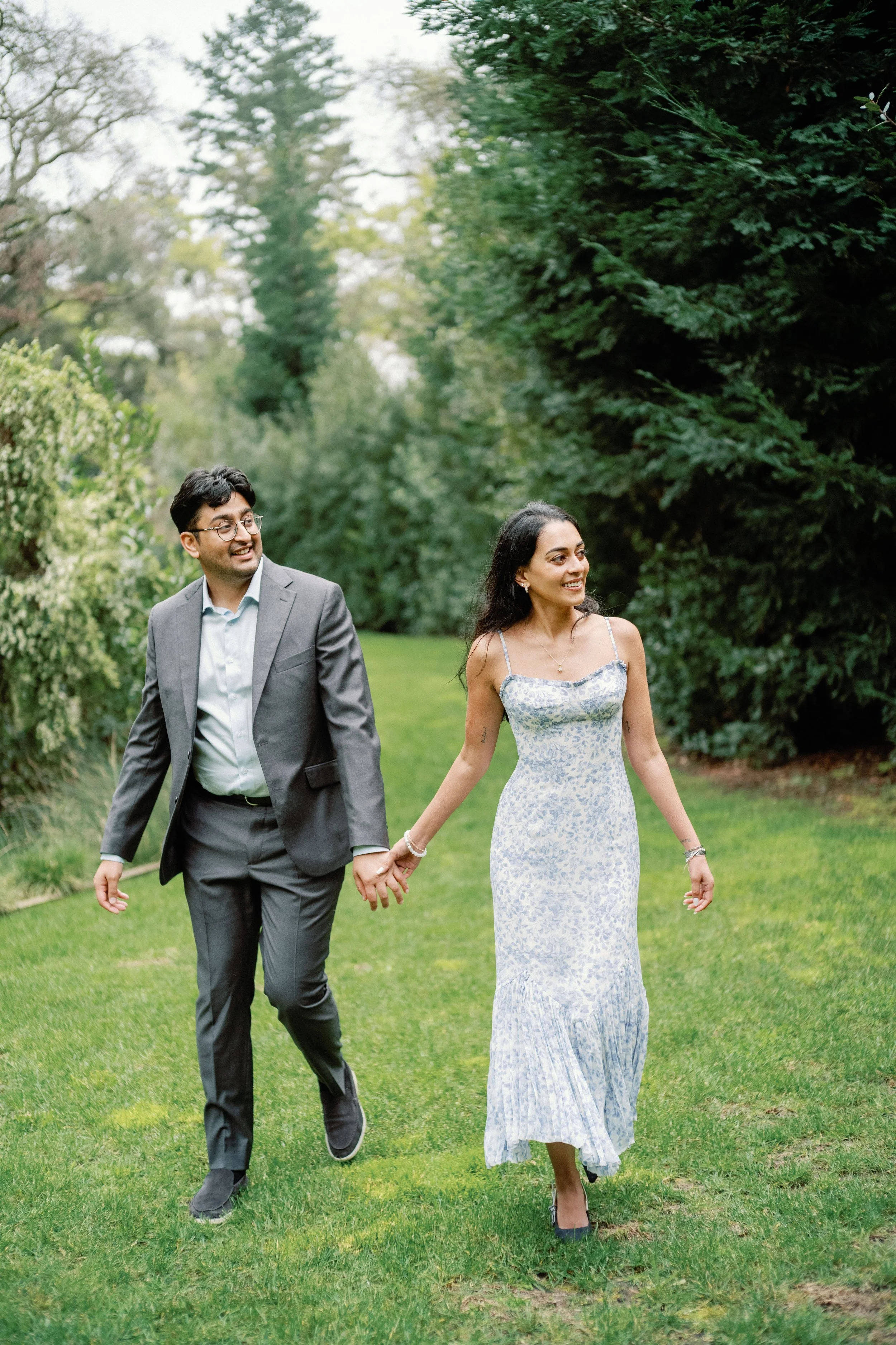 A couple walking hand in hand on a grassy path, surrounded by green trees and bushes, smiling and enjoying a sunny day outdoors.