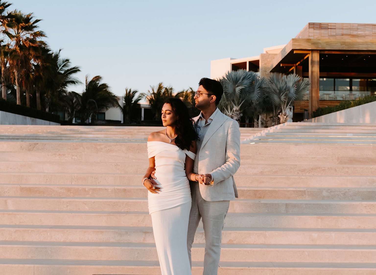 A couple dressed in elegant clothing standing on a set of stairs outdoors during sunset, with palm trees and modern buildings in the background.