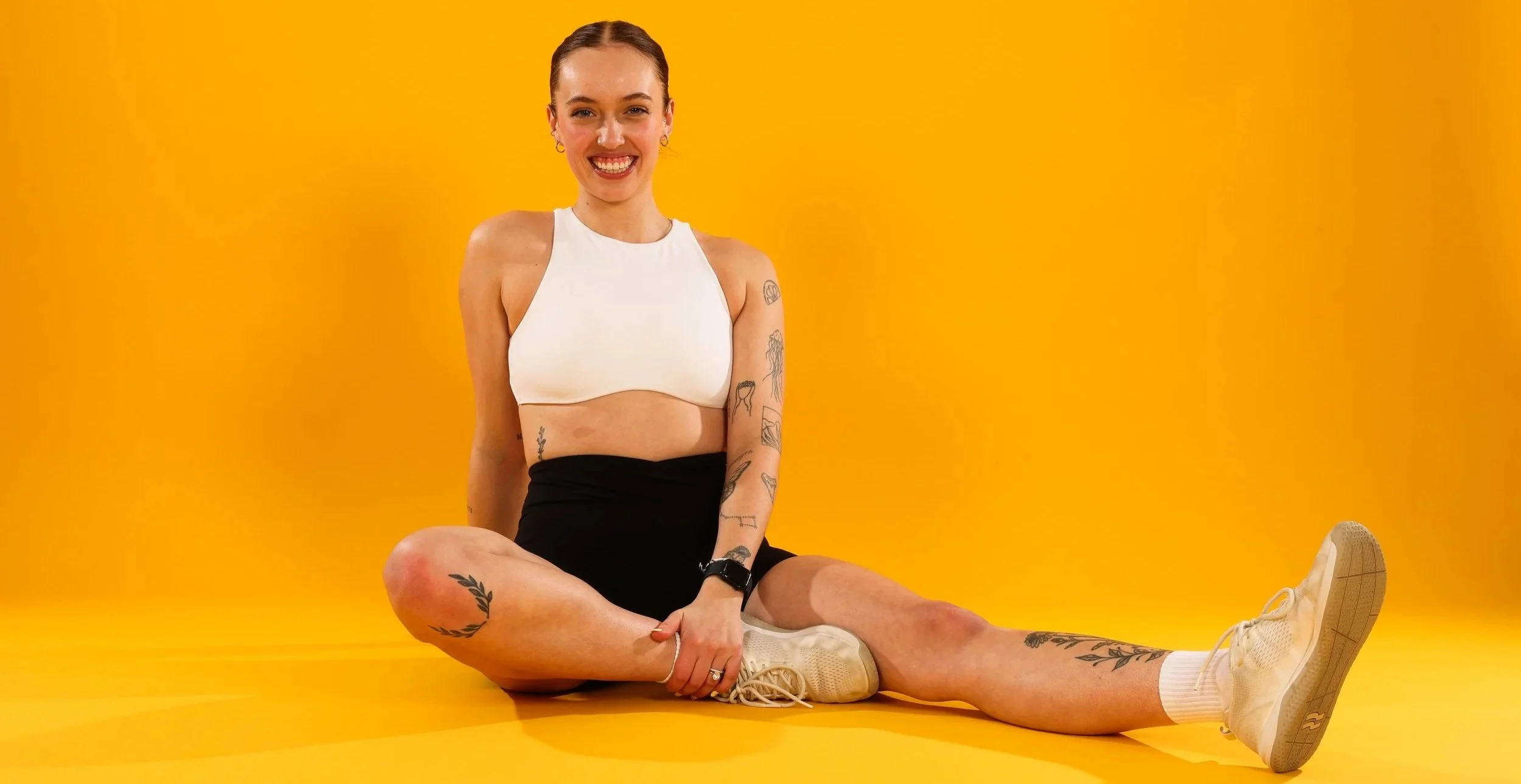 A young woman with tattoos sits on a yellow background, smiling, wearing a white sleeveless crop top and black shorts, with her legs extended and yellow sneakers.