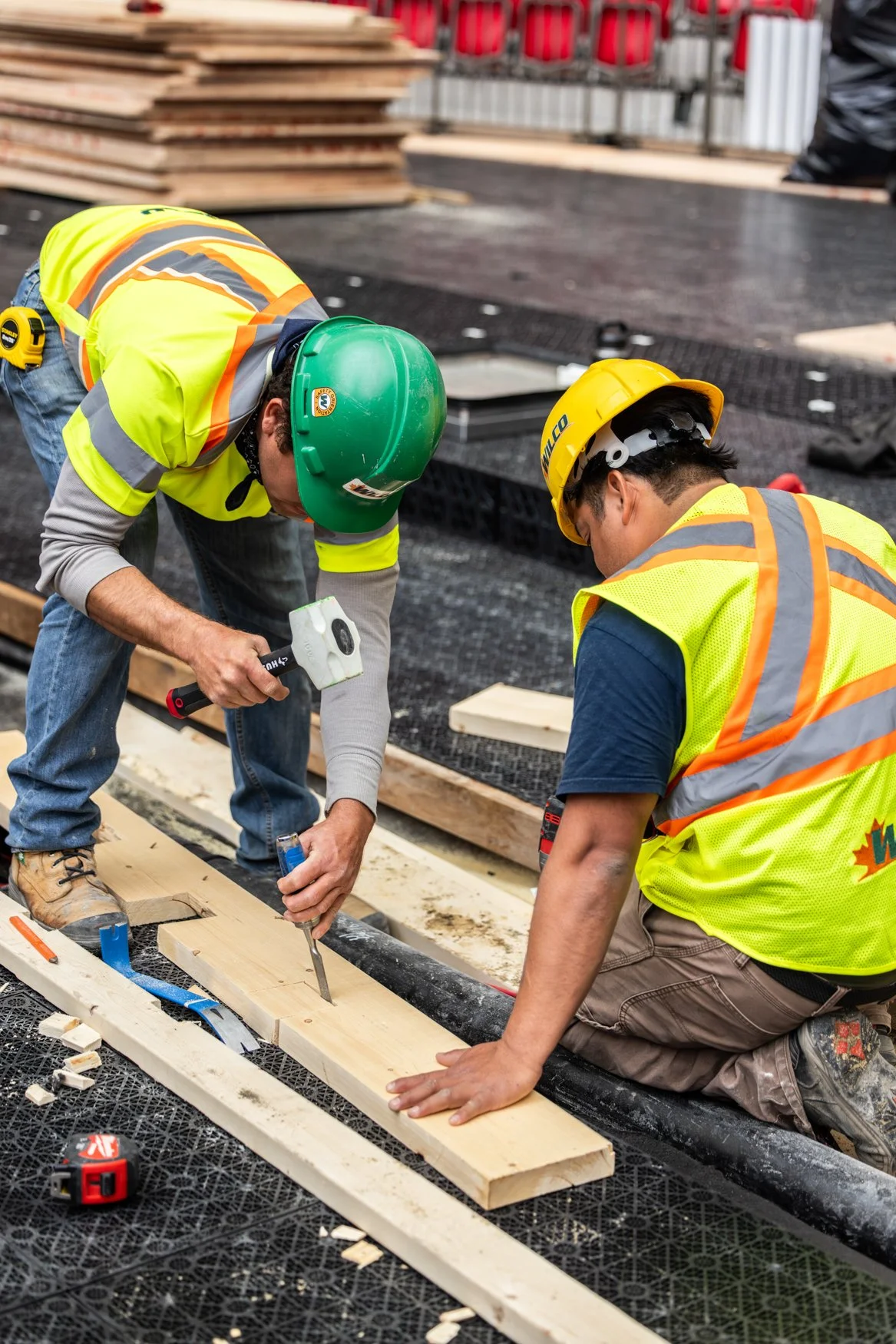 Two construction workers in safety vests and helmets kneeling on a building site, working with wooden planks and tools.