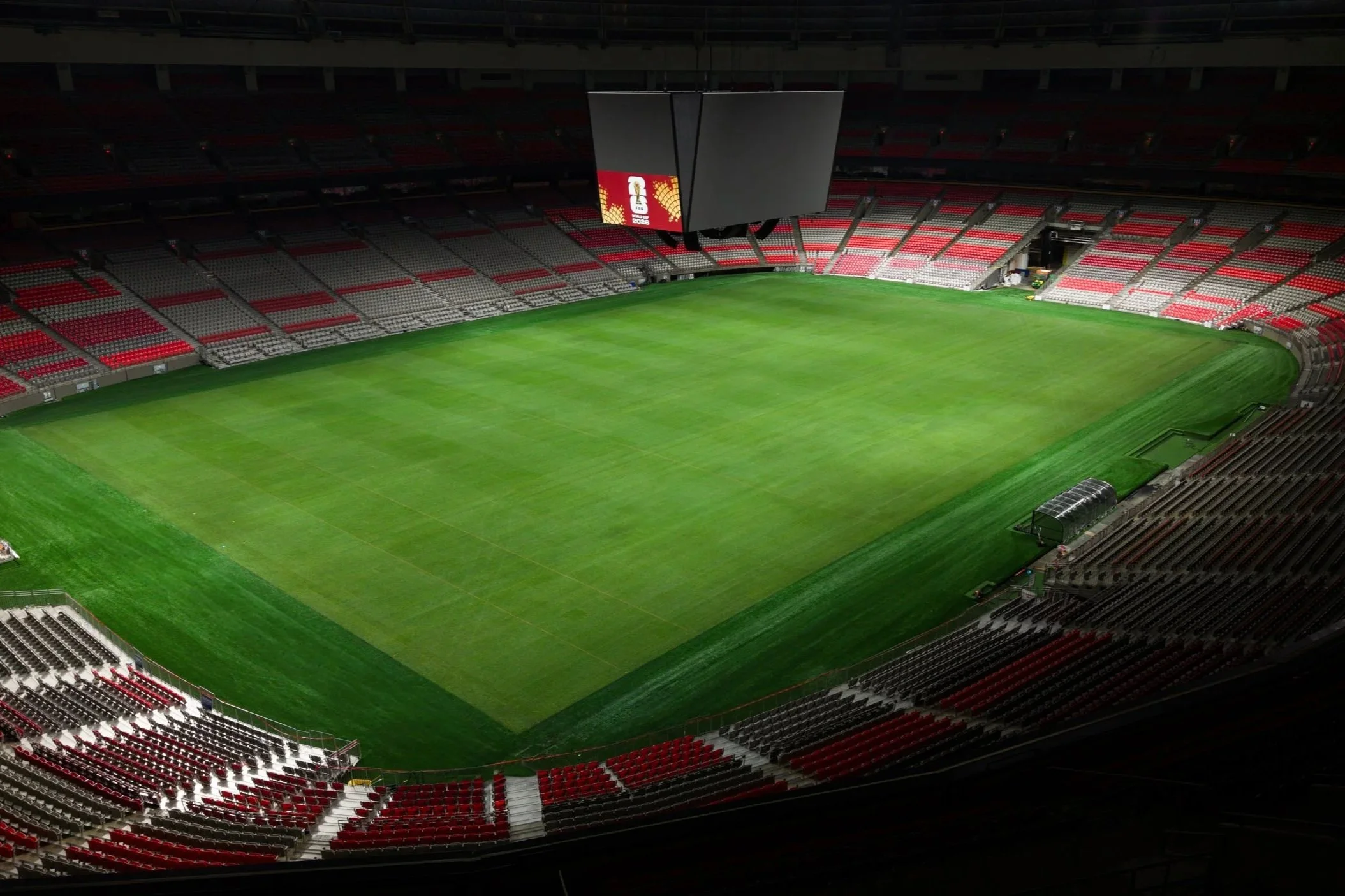 Empty soccer stadium with green field, red and white seating, and large overhead scoreboard.