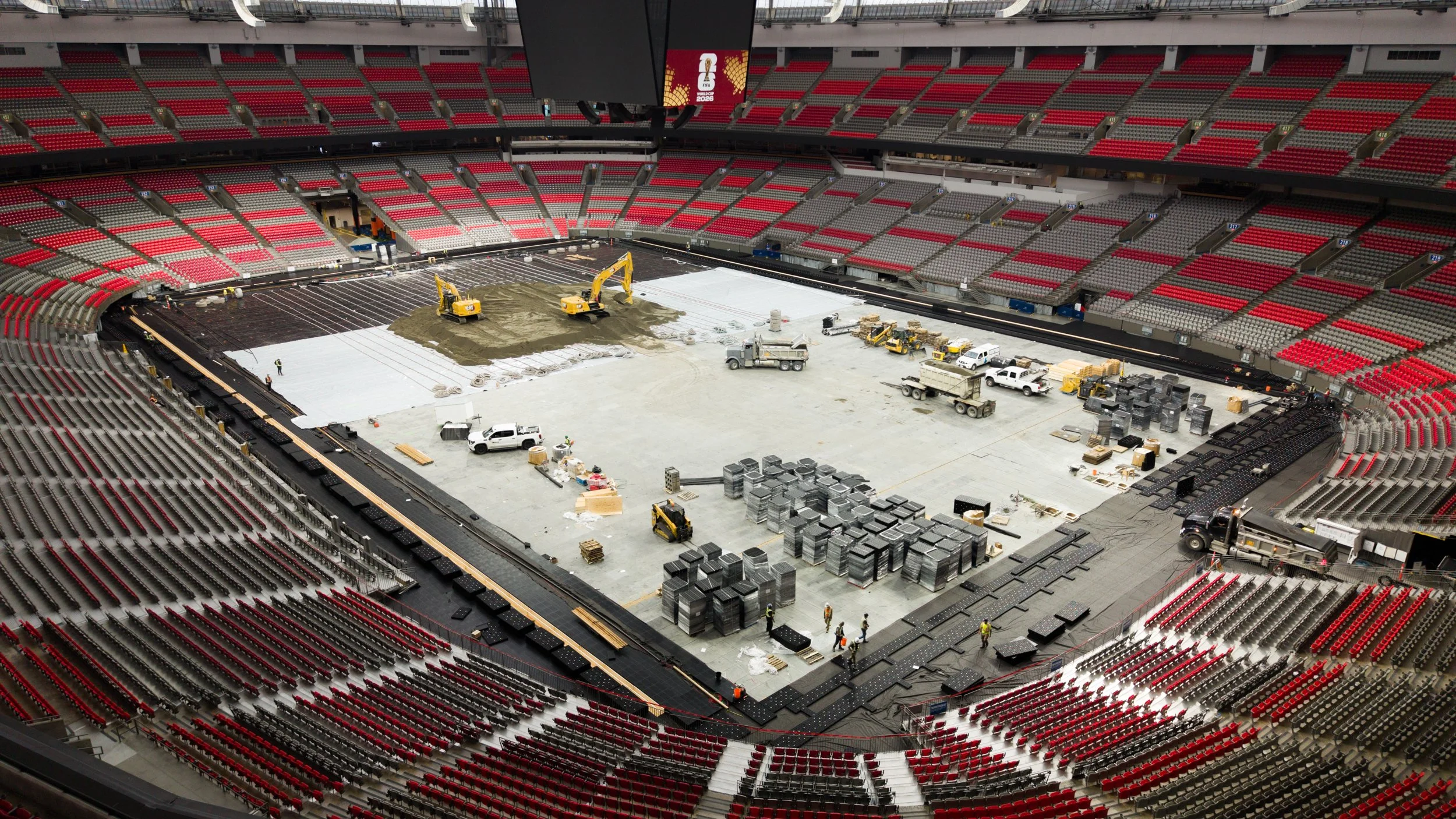 Inside a large stadium under construction, with red and gray seating surrounding the field, construction vehicles and materials in the center, and a large screen displaying a logo above.