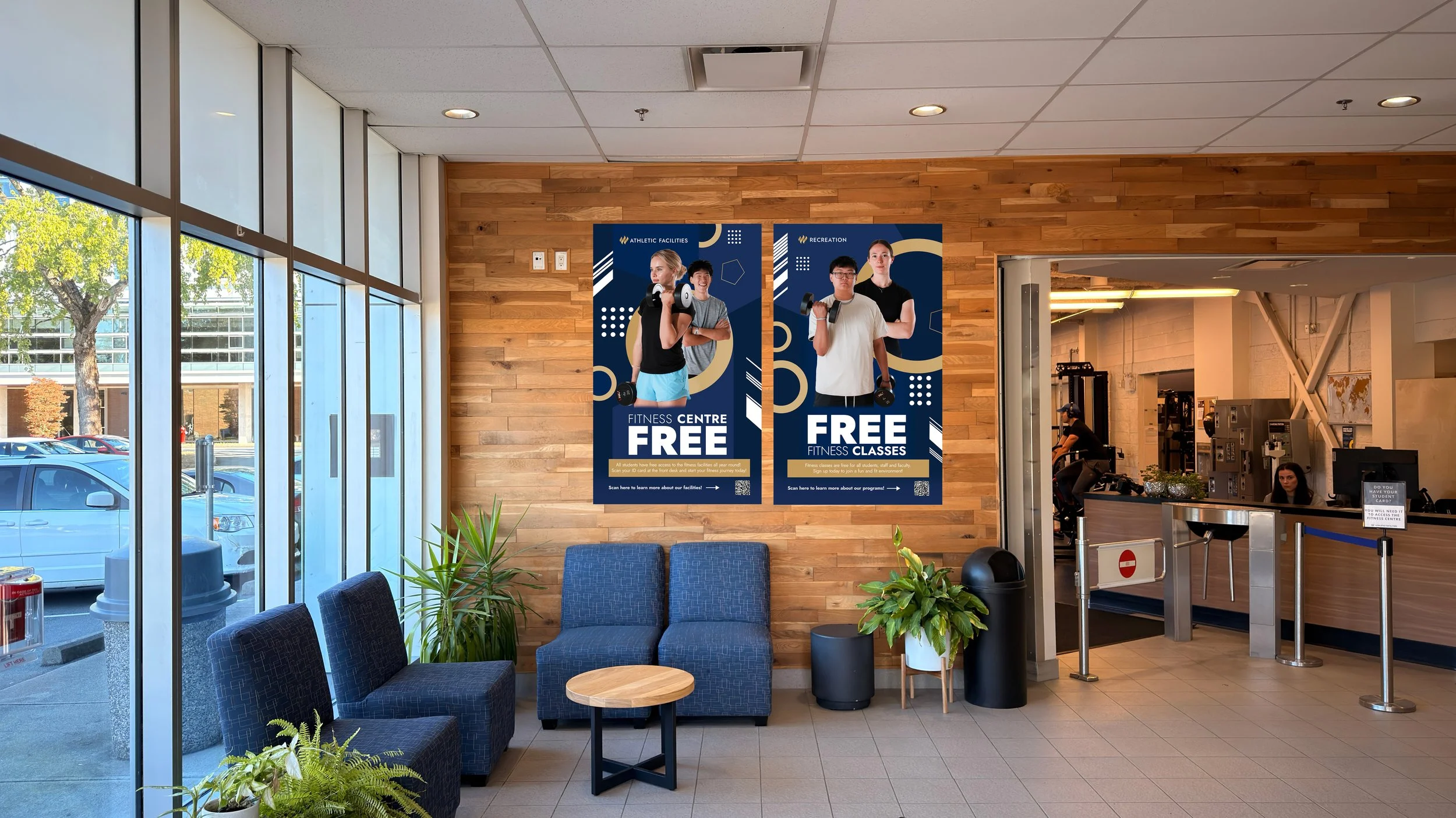 Indoor view of a fitness center reception area with promotional posters on a wooden wall, seating with blue chairs and plants, large windows showing parked cars outside, and a front desk area with staff.