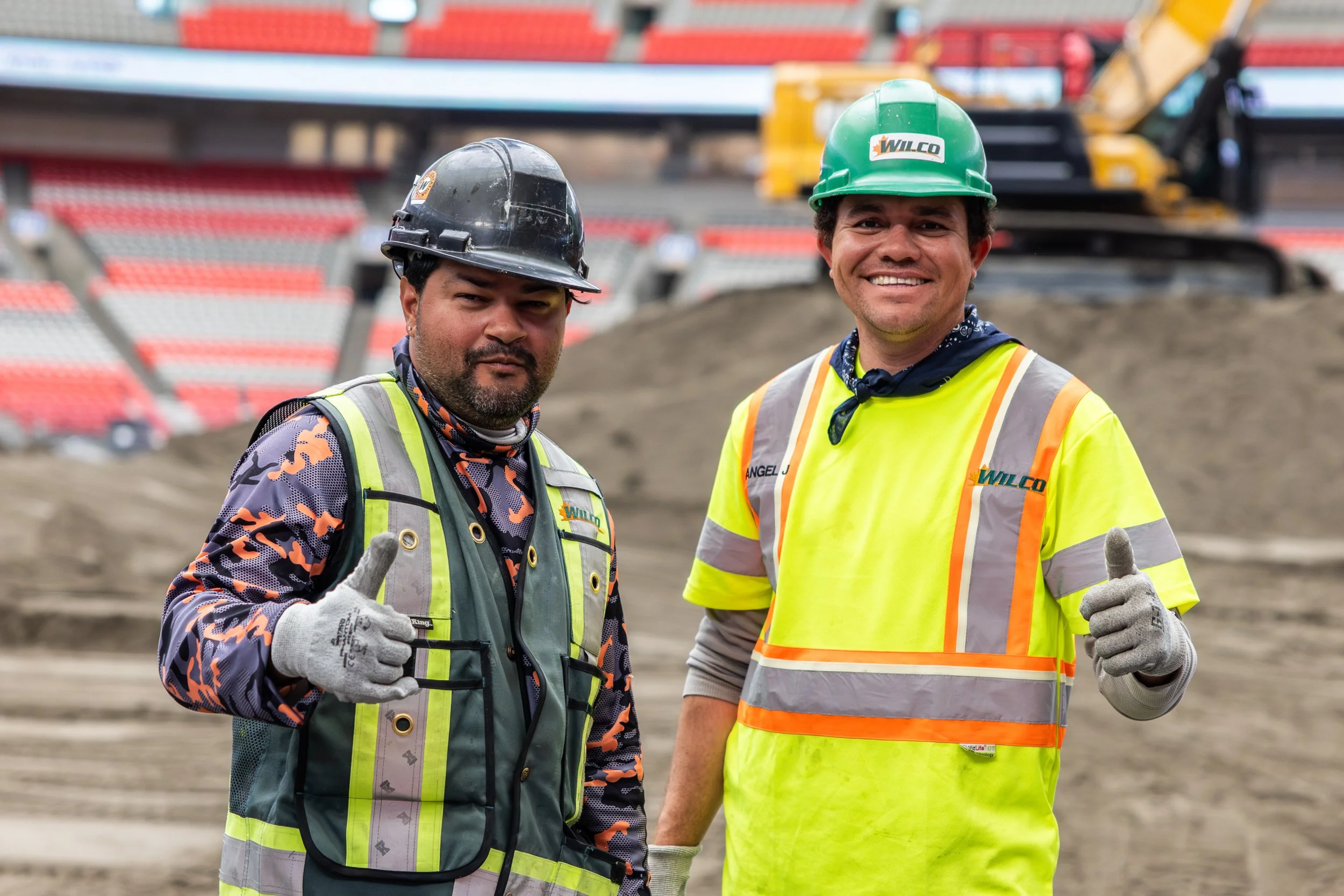 Two construction workers standing at a construction site, smiling and giving thumbs up. One is wearing a black helmet and a patterned shirt with safety vest, the other is wearing a green helmet and a yellow safety vest.