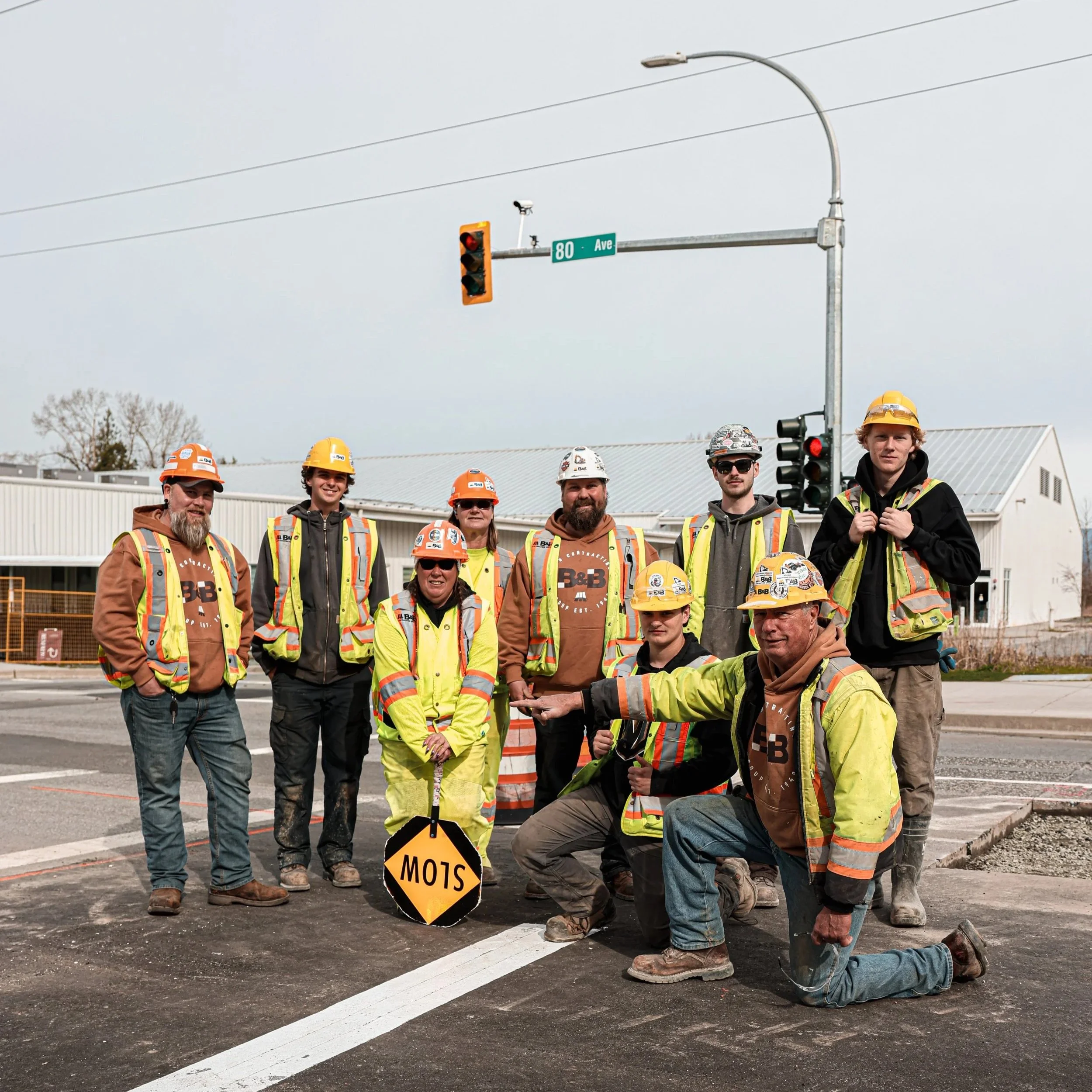 Group of construction workers and engineers standing and kneeling on a newly paved road, with a traffic light, a warning sign, and a building in the background.