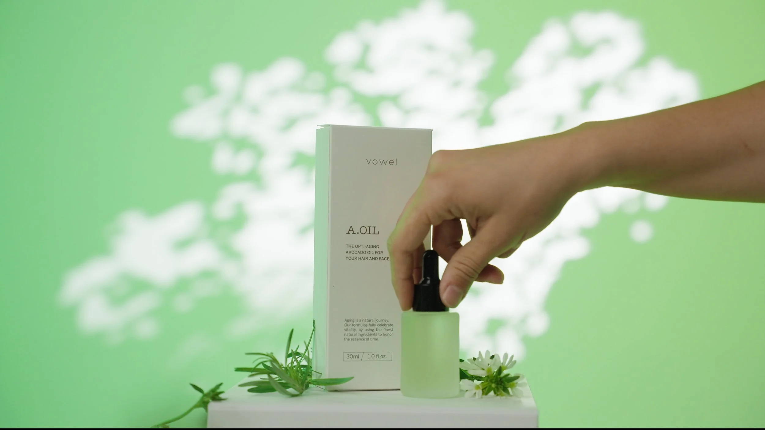 A hand holding a dropper above a frosted glass bottle of avocado oil labeled 'A.OIL' with a white box labeled 'vowel' in the background, all placed on a white surface with green leaves and white flowers, against a green background with white cloud-like shadows.