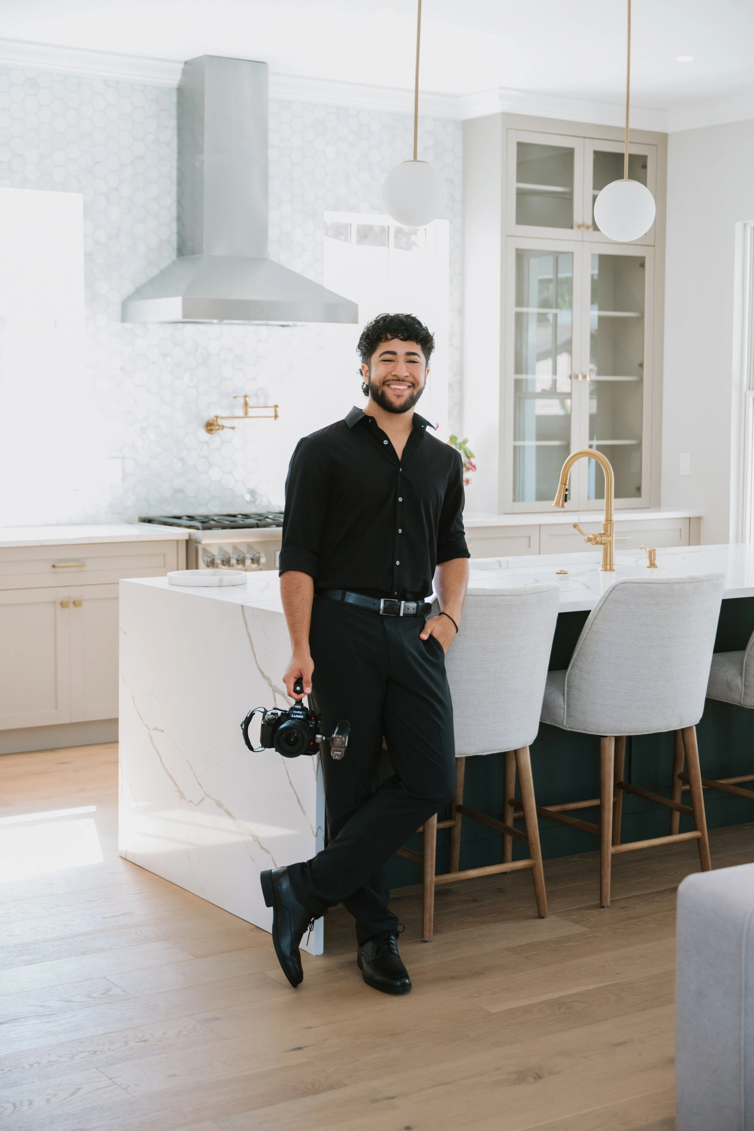Young man with dark curly hair and a beard, wearing a black shirt and dress pants, smiling while holding a camera, standing in a modern kitchen with white countertops, gold fixtures, and bar stools.