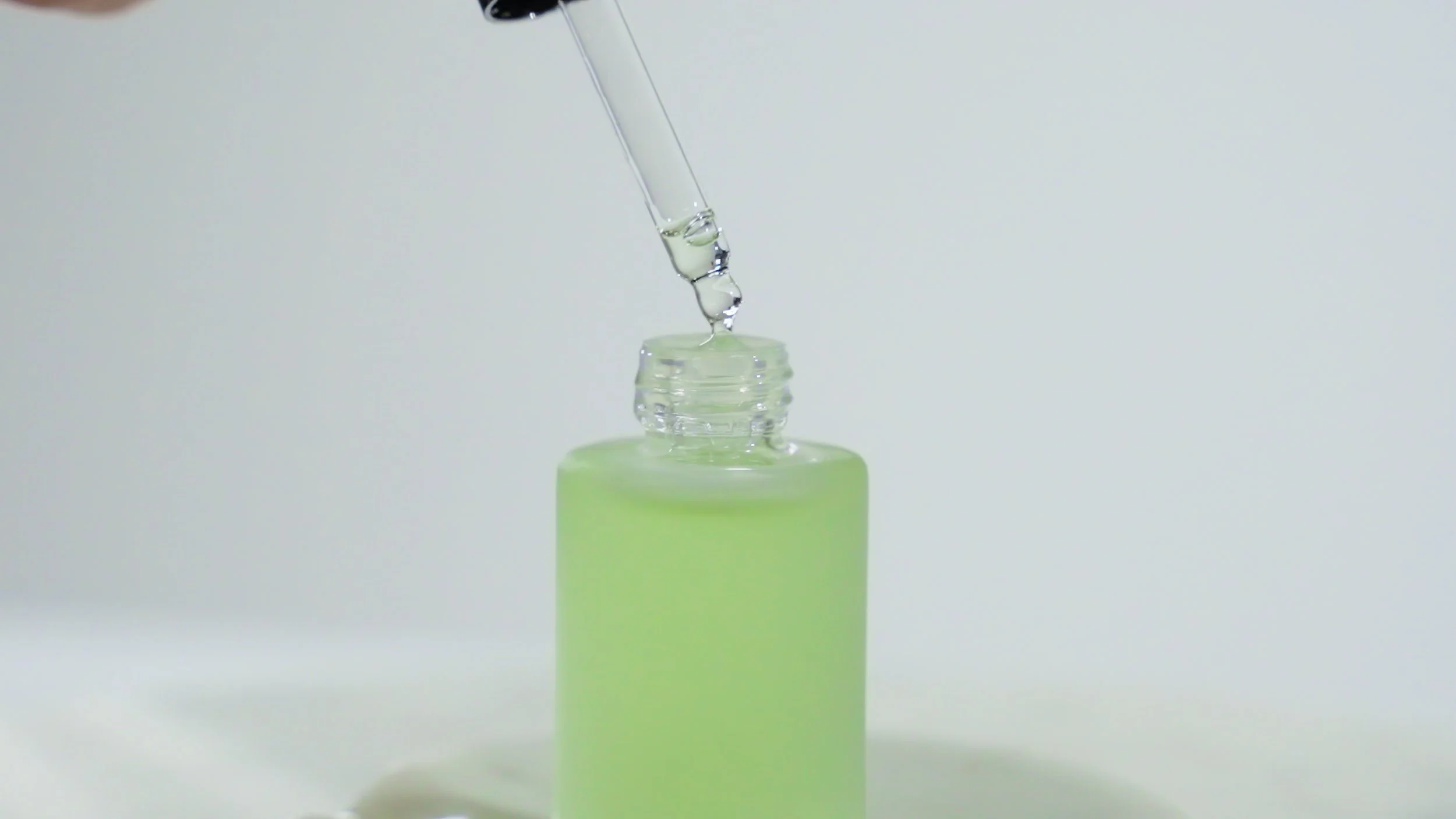 A clear dropper is releasing a drop of liquid into a light green bottle with a white background.