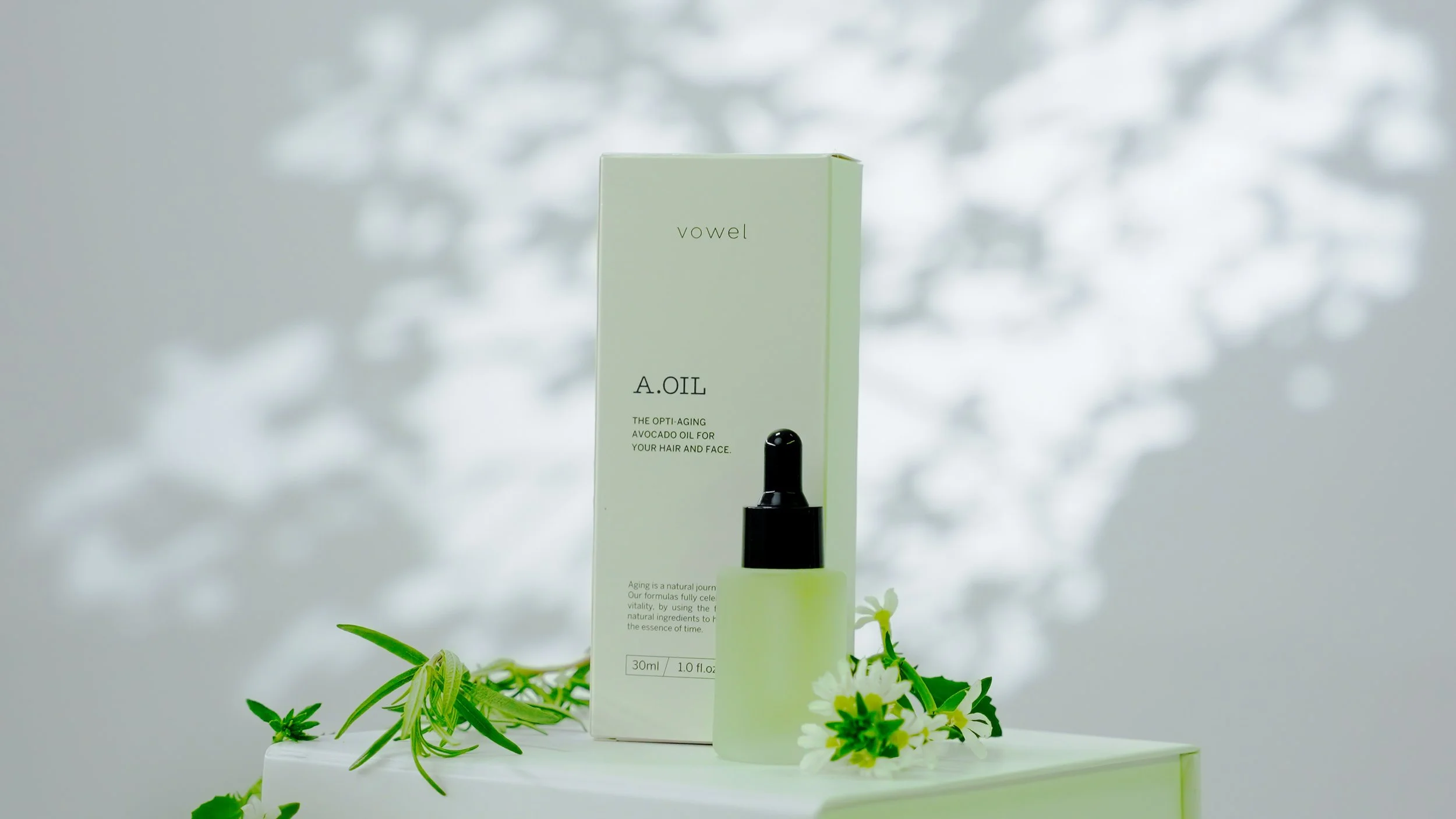 A white box labeled 'vowel A.OIL' with a small bottle of oil beside it, decorated with green leaves and white flowers, on a white surface with soft shaded background.