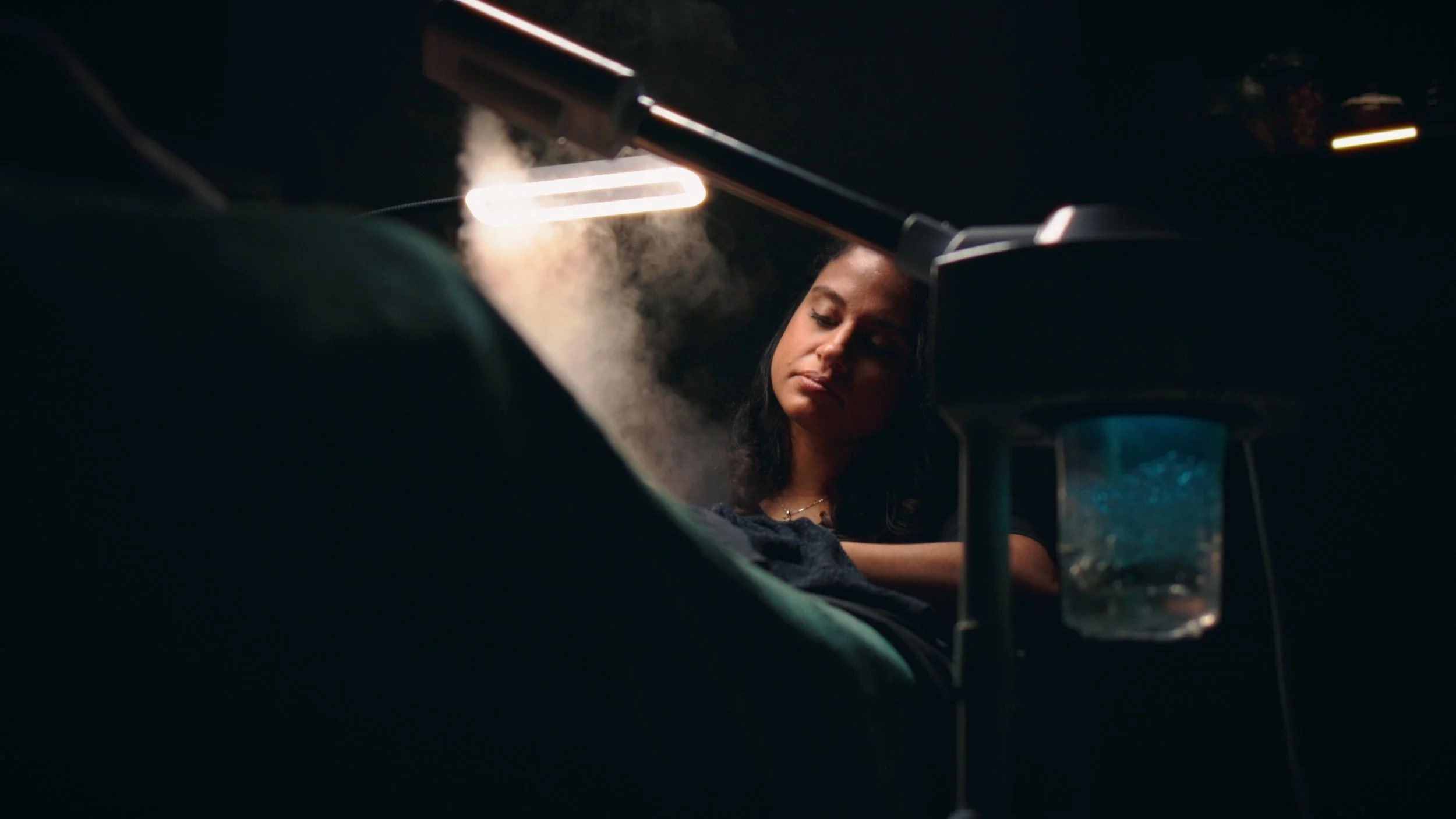 A woman is lying in a dimly lit room, with a neon light overhead and a steam vapor around her, appearing to be in a medical or relaxation setting.