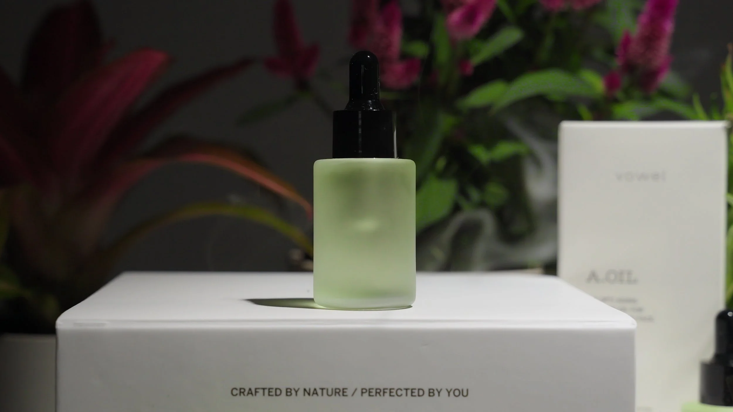 A small frosted glass bottle with a black dropper cap, placed on a white box with the phrase "Crafted by nature / perfected by you" beneath. In the background, there are pink and green plants and a white box labeled "vowel."