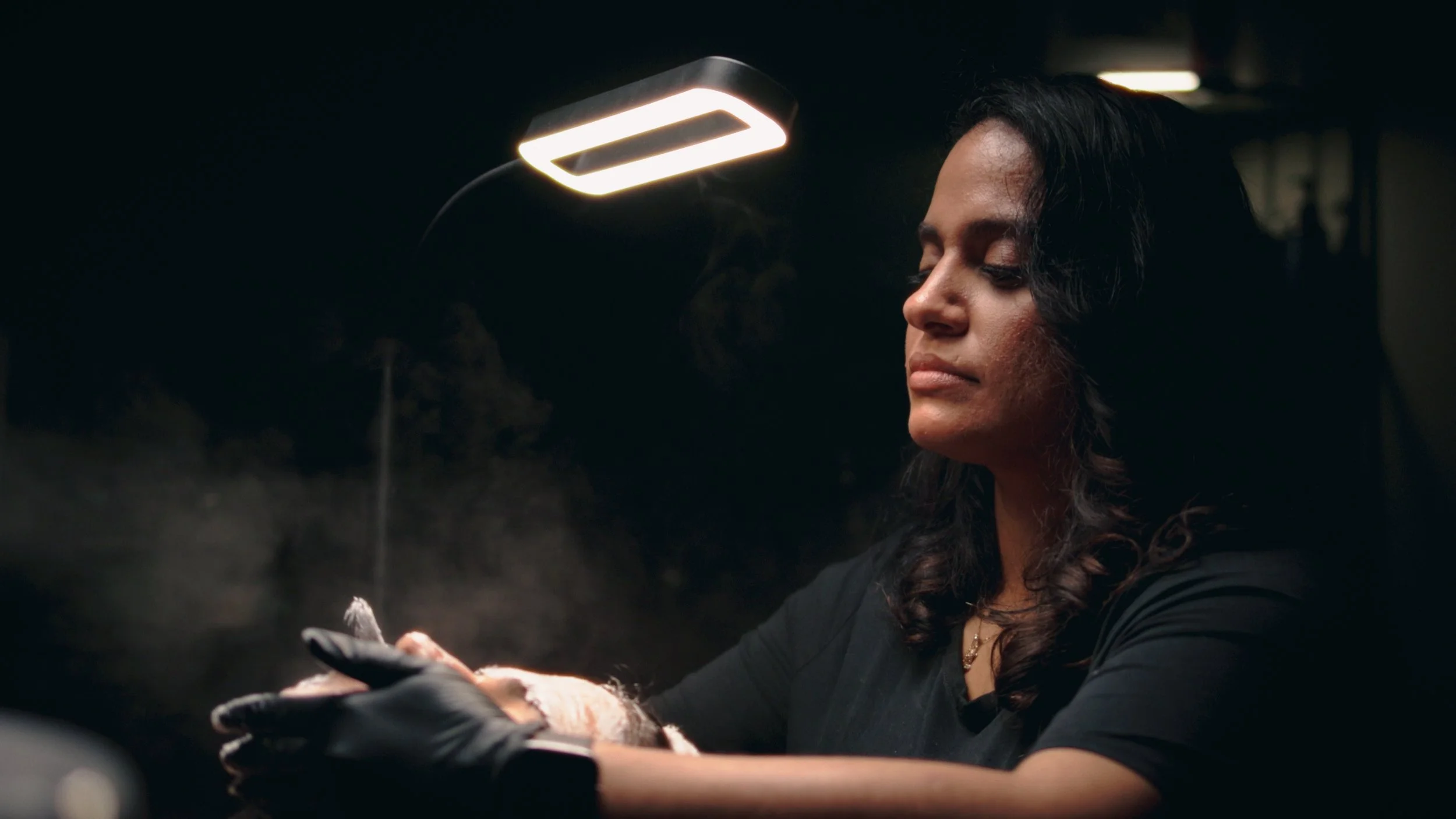 A woman with dark hair, wearing a black shirt and black gloves, is performing a task in a dark room illuminated by a bright overhead lamp.