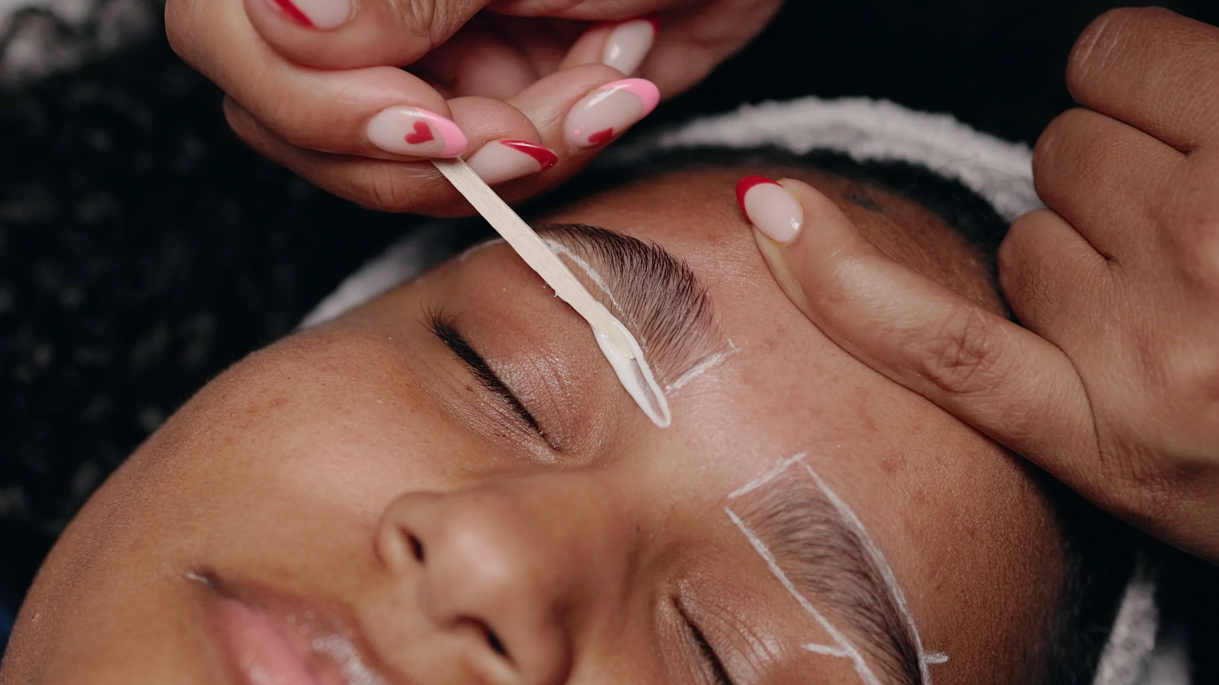 A person receiving an eyebrow waxing or threading treatment with white markings on the eyebrows, while a technician applies wax or threading paste.