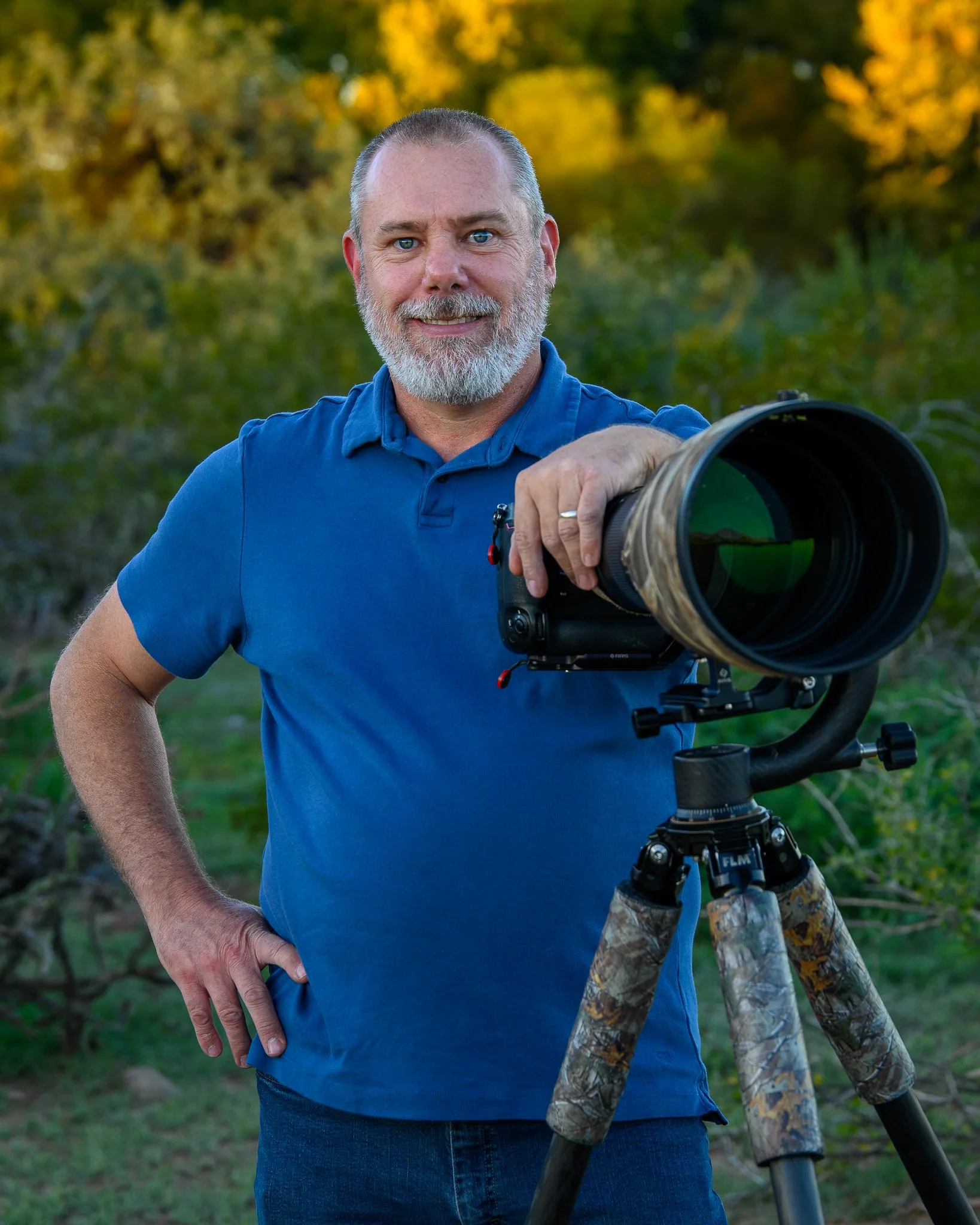 A man with a gray beard and short hair standing outdoors, smiling, wearing a blue polo shirt, holding a large camera with a long lens mounted on a tripod, against a background of trees with fall foliage.